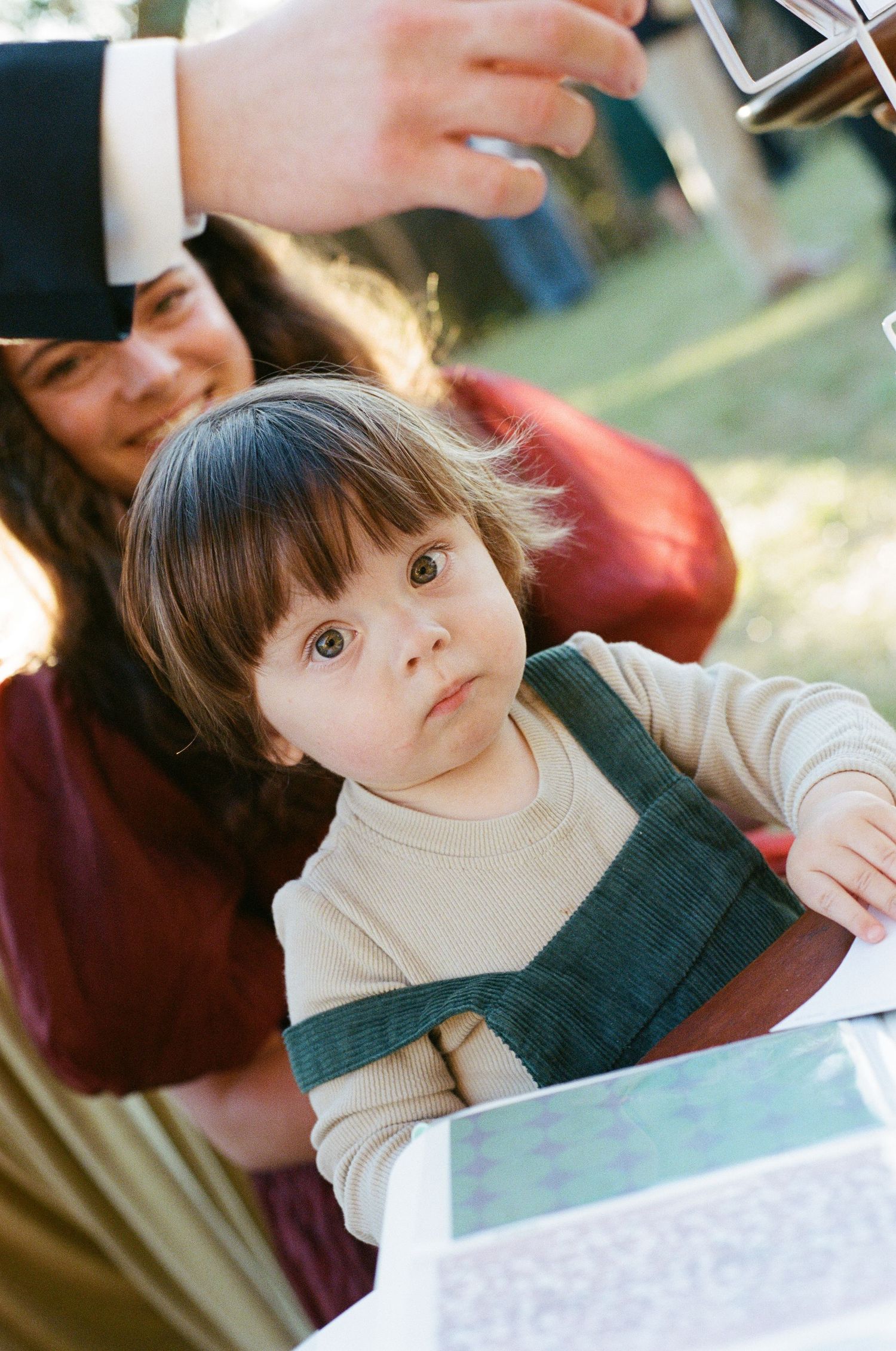 Adorable toddler with wide brown eyes sitting with parents at an outdoor wedding reception table.