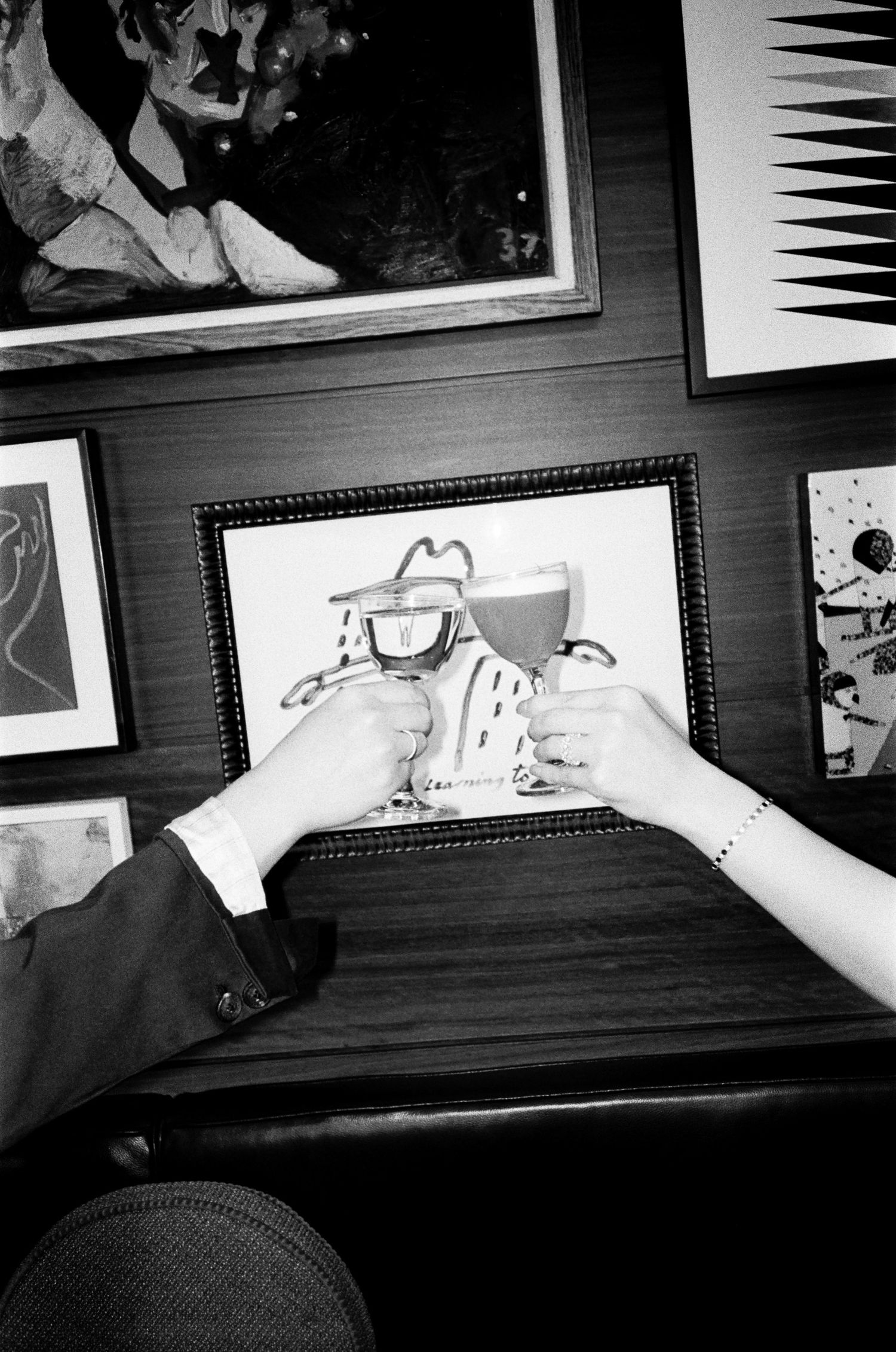 Two people clink wine glasses in a toast at a wooden booth, with framed artwork covering the wall behind them in a black and white photo.