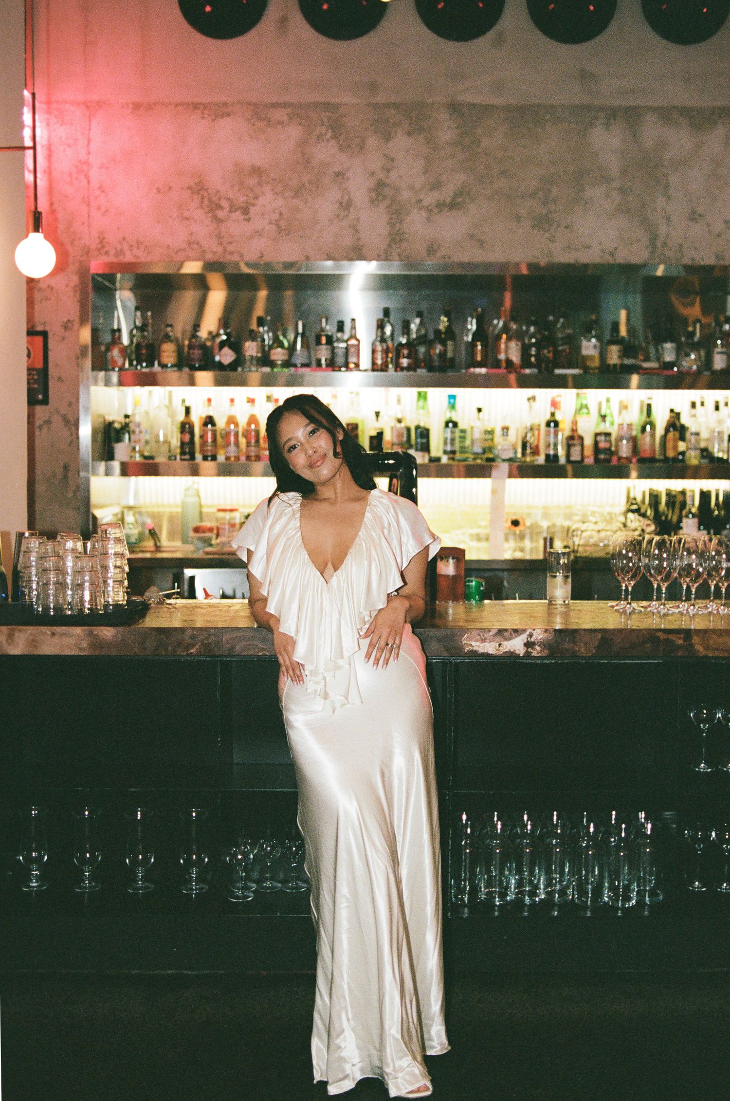Woman in a flowing white satin gown with a plunging neckline poses in front of a stylish bar with backlit liquor bottles and wine glasses.
