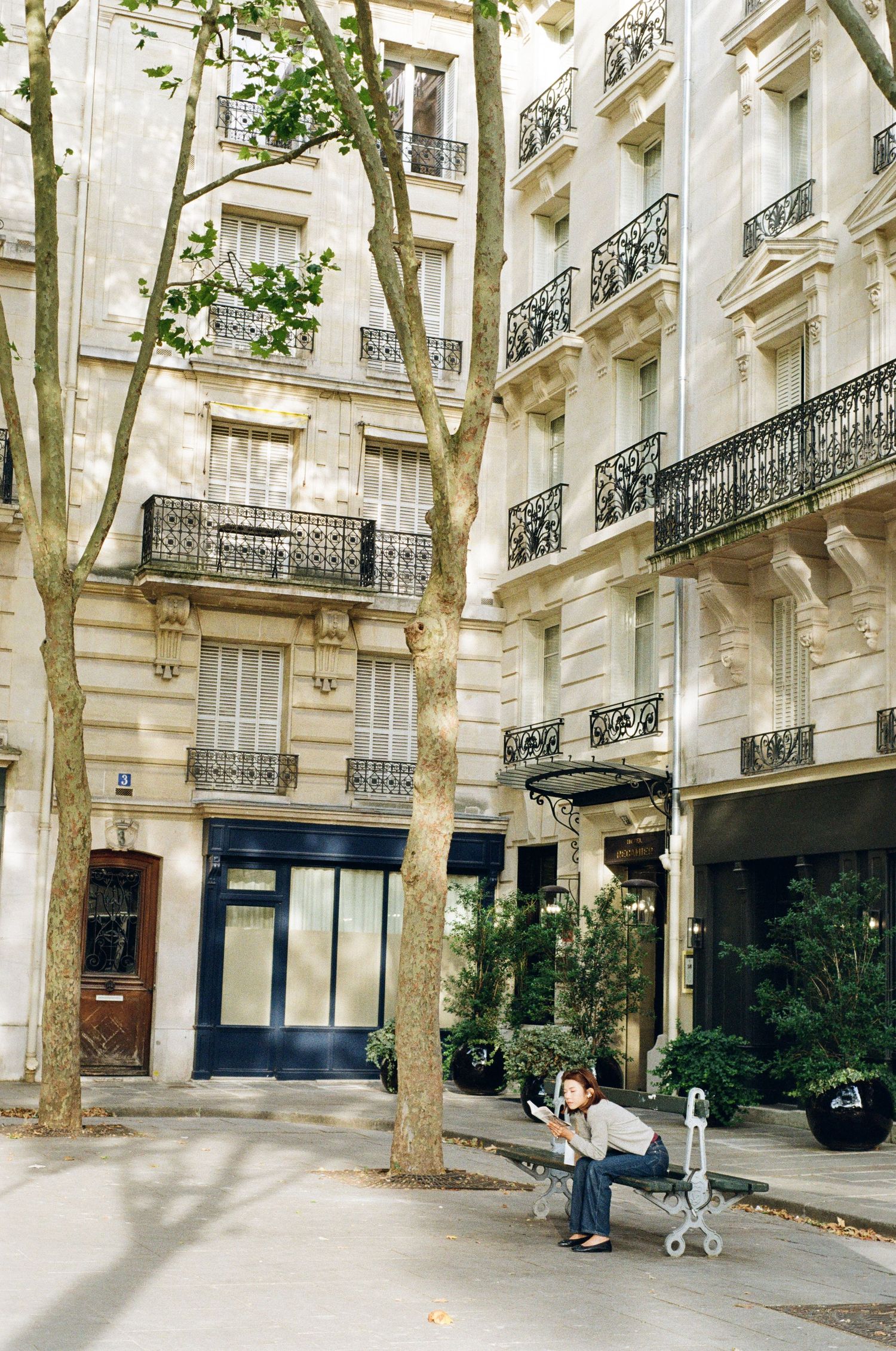 Woman sitting on a bench reading beneath a tree on a sunlit Parisian street lined with Haussmann buildings and wrought-iron balconies.
