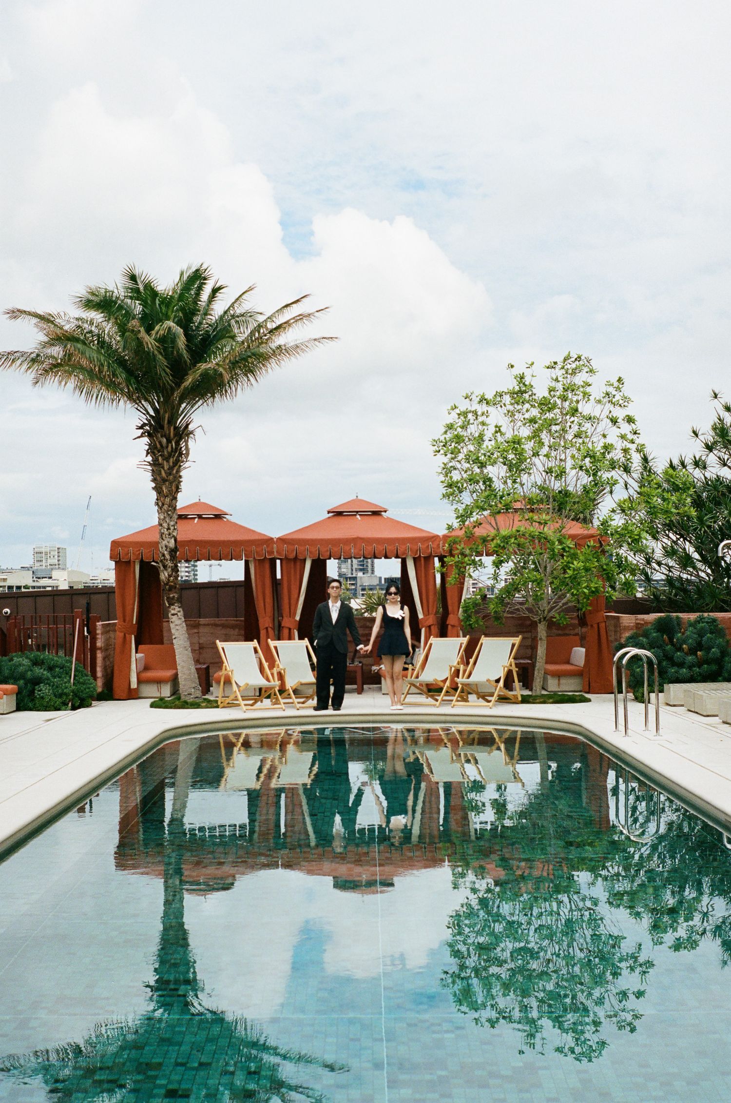 Couple stands between orange poolside cabanas and lounge chairs beside a rooftop pool with palm trees reflected in the water.