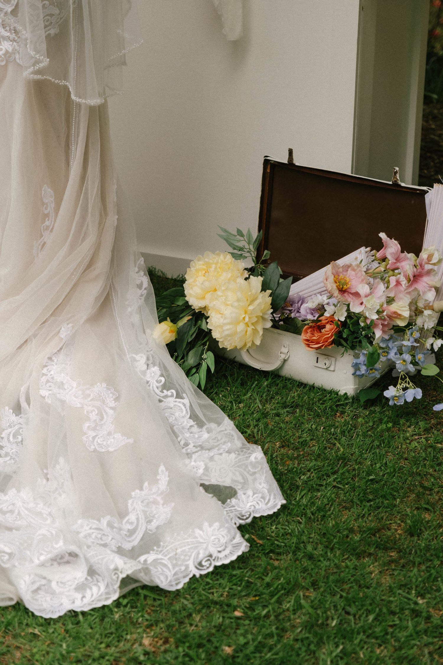 Elegant white lace wedding dress hems and colorful floral arrangements displayed on green grass outdoors.
