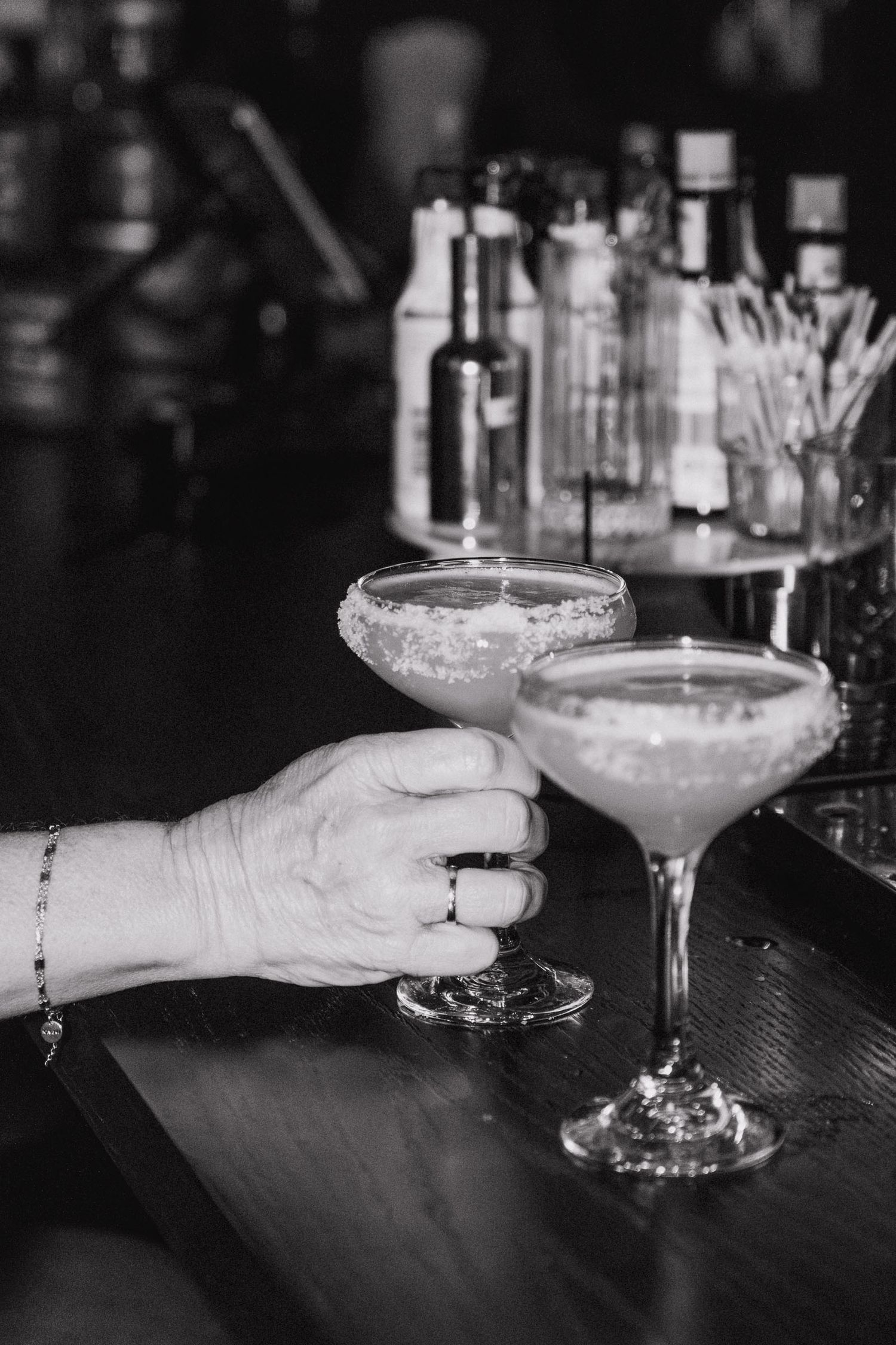 Black and white photo series showing hands holding elegant cocktail glasses with frothy drinks on a dark bar counter.