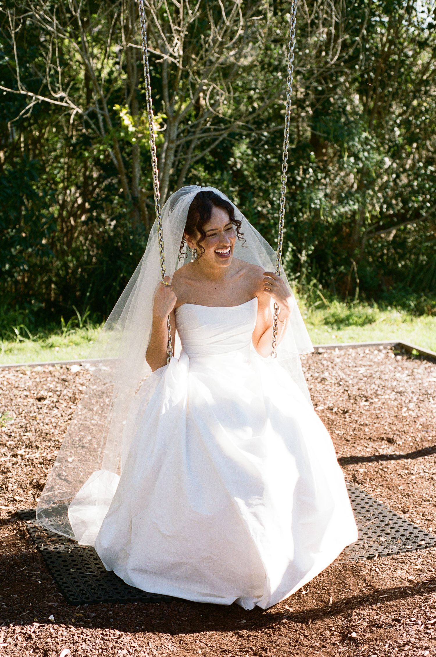 Smiling bride in a strapless white wedding gown and sheer veil laughs while sitting on a chain swing outdoors surrounded by green trees.