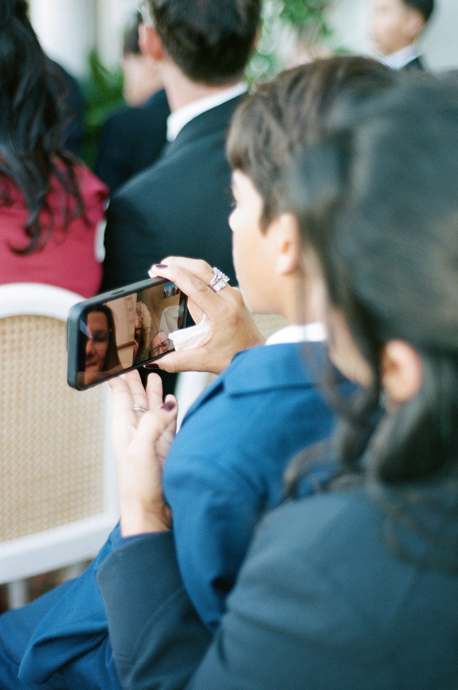 Wedding guest in a blue blazer holds up a smartphone to record a ceremony while seated among other formally dressed attendees.