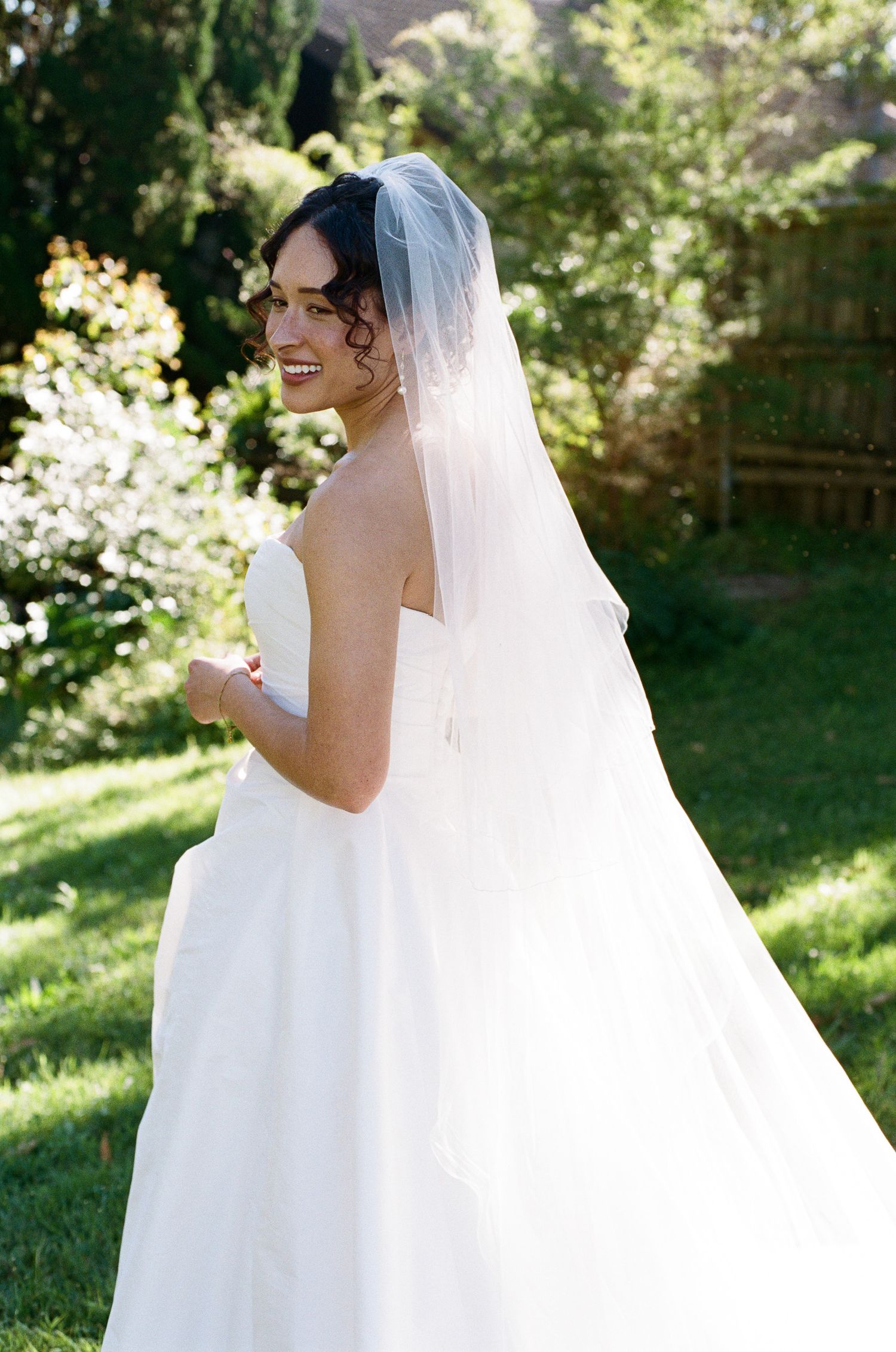 Smiling bride in a strapless white wedding gown and long sheer veil glancing over her shoulder in a sunlit garden.