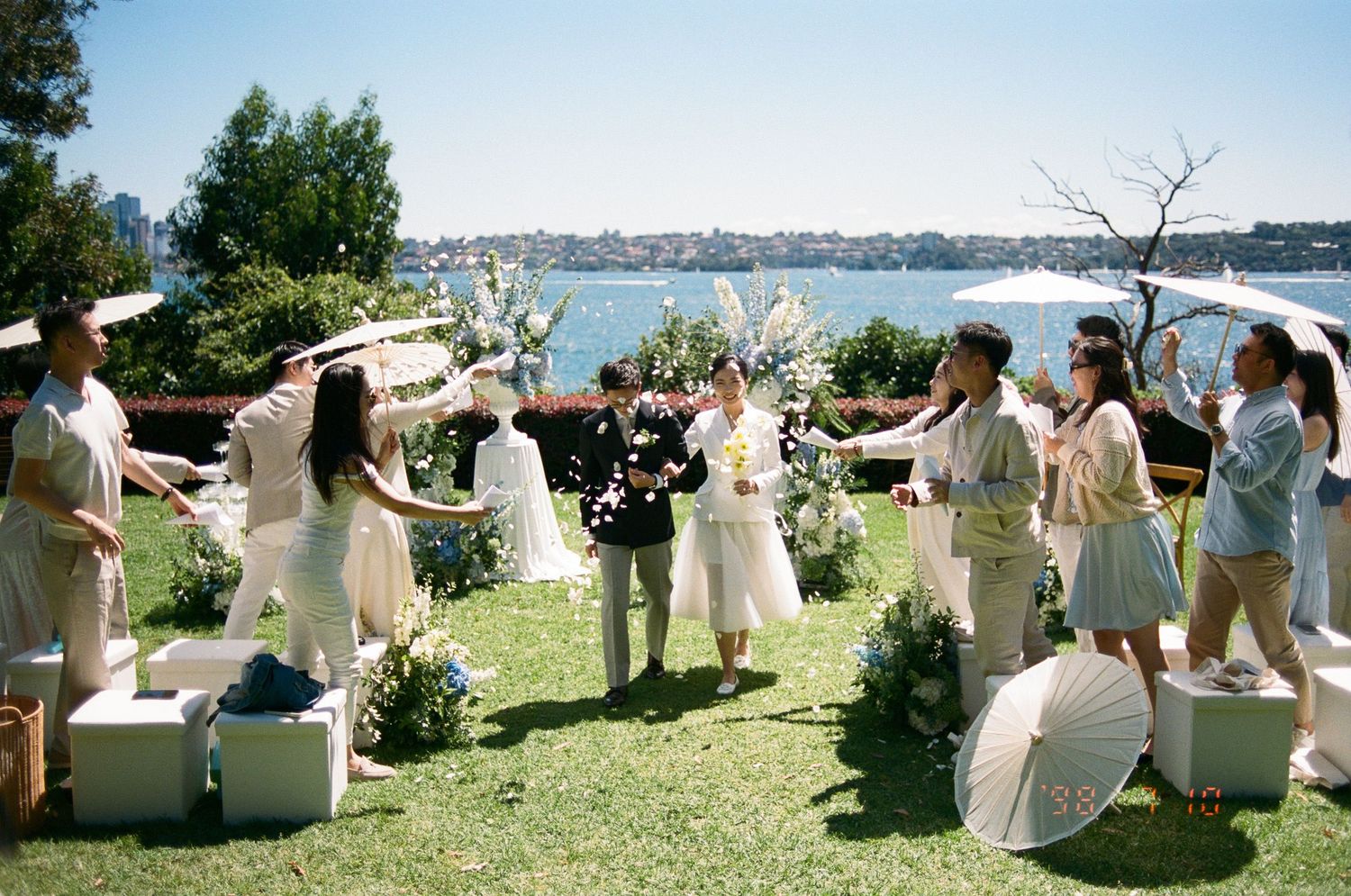Bride and groom walk down the aisle as guests in white and neutral outfits toss petals at an outdoor waterfront wedding ceremony.