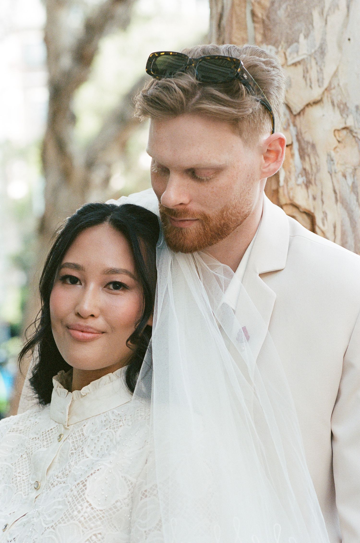 A bride in a white lace high-collar dress and veil leans against a groom in a cream suit with sunglasses on his head, beside a tree.