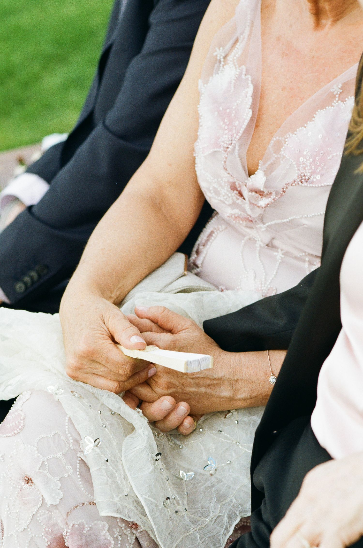 Seated wedding guests hold hands, one in a lace-trimmed blush dress clasping a folded paper, beside a person in a dark suit.