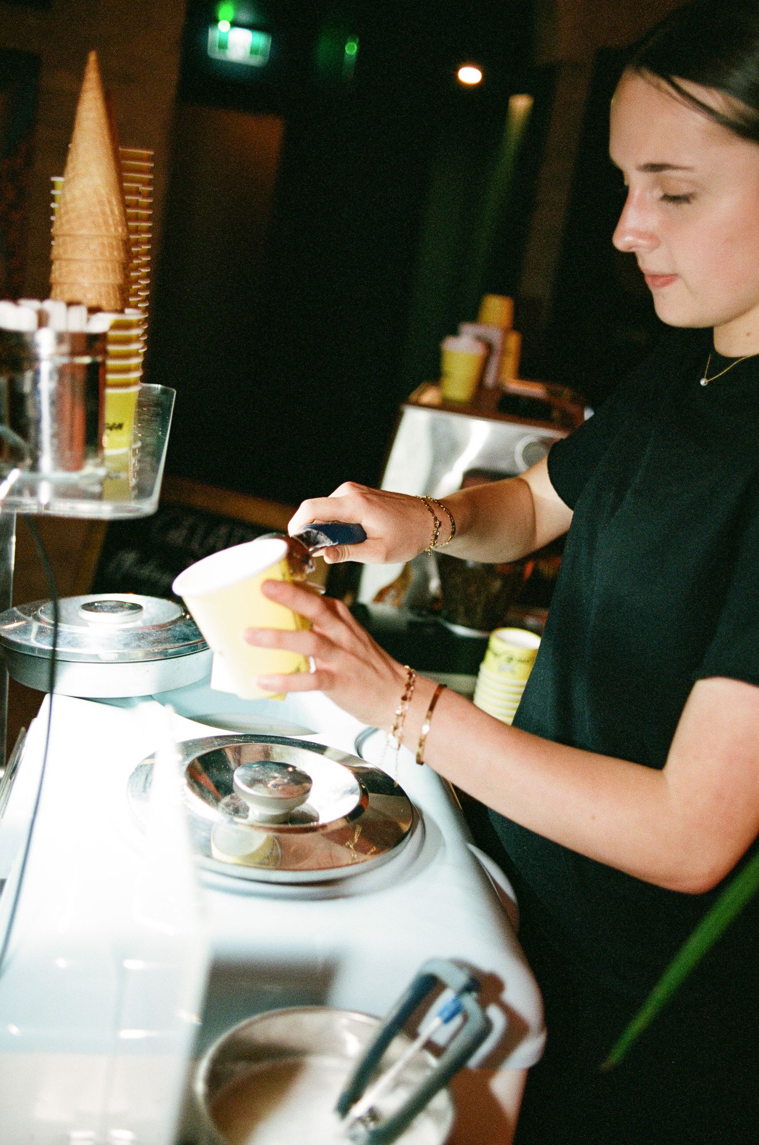 A woman in a black shirt scoops ice cream into a cup at a gelato counter, with a stack of waffle cones beside her.