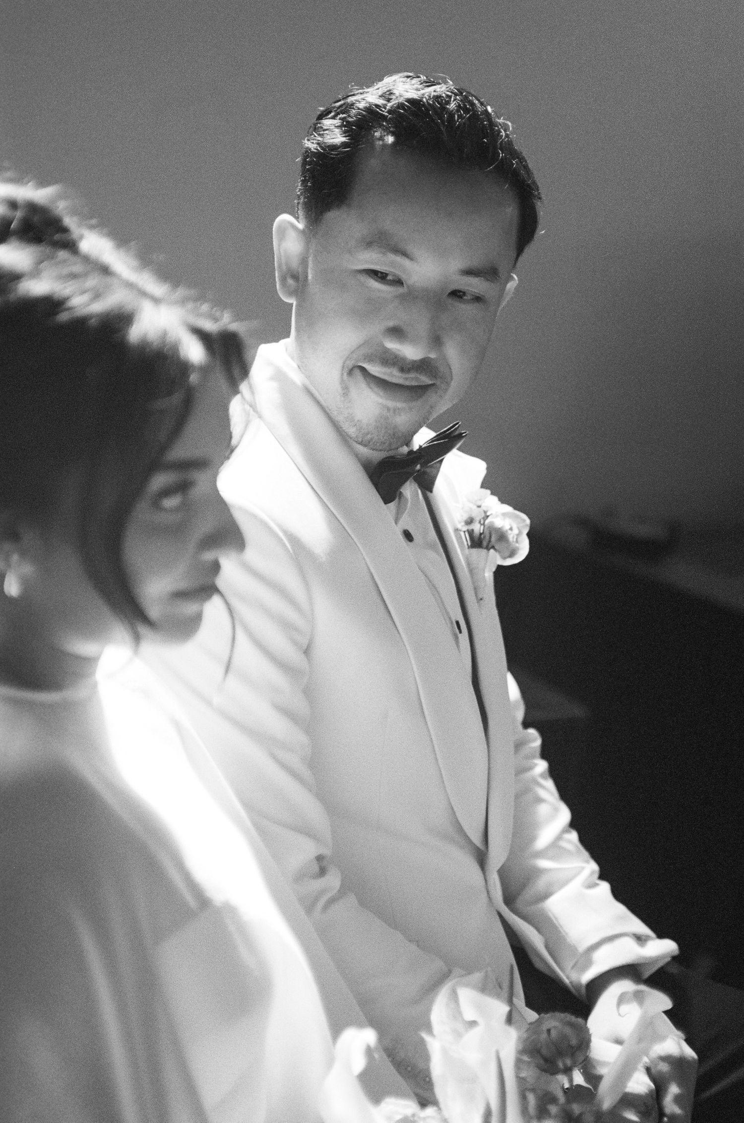 Black and white portrait of a smiling groom in a white tuxedo jacket, bow tie and boutonniere standing beside a bride in soft window light.