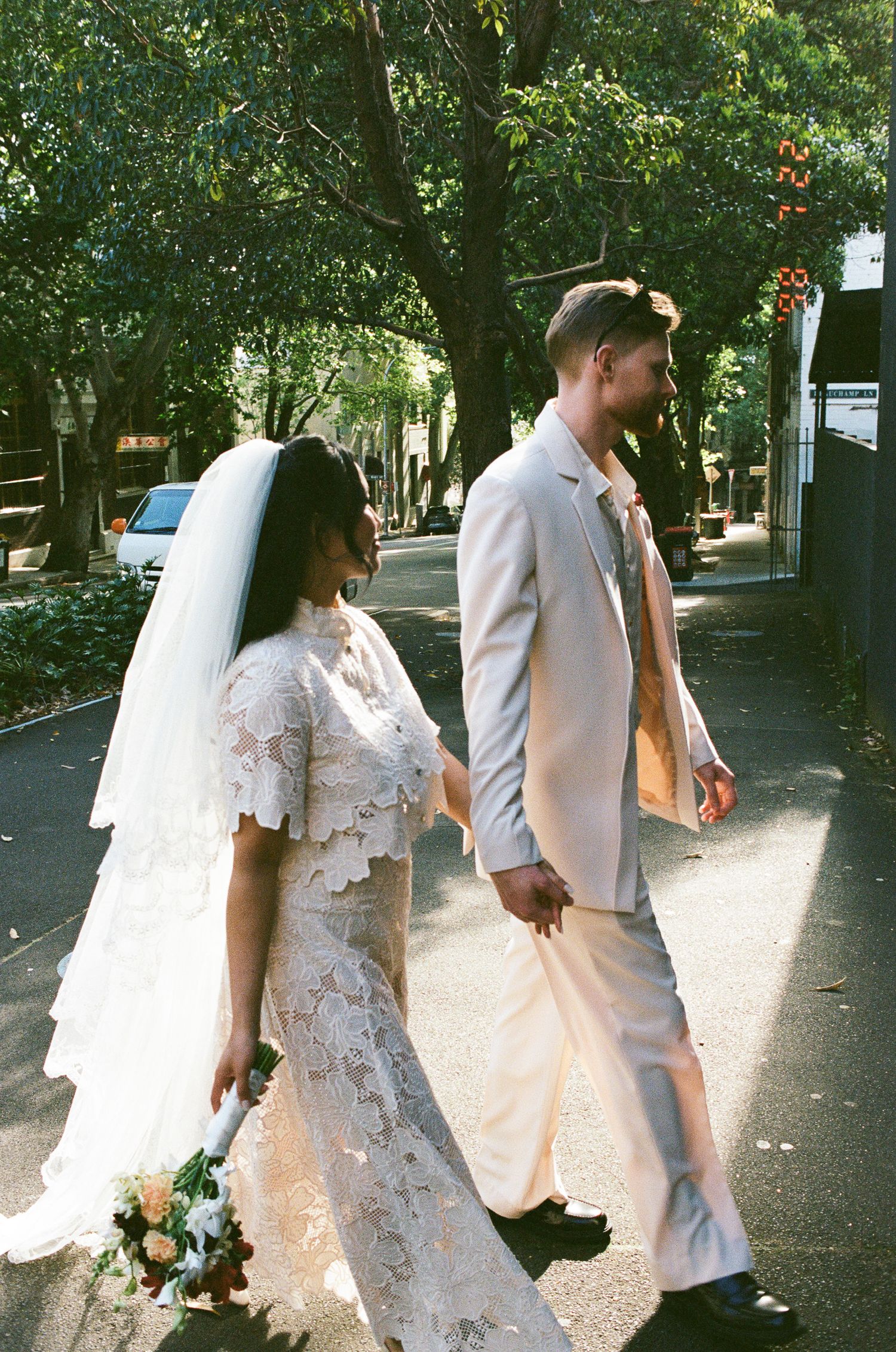Bride in a lace gown and veil holding a bouquet walks hand in hand with a groom in a cream suit down a tree-lined street.