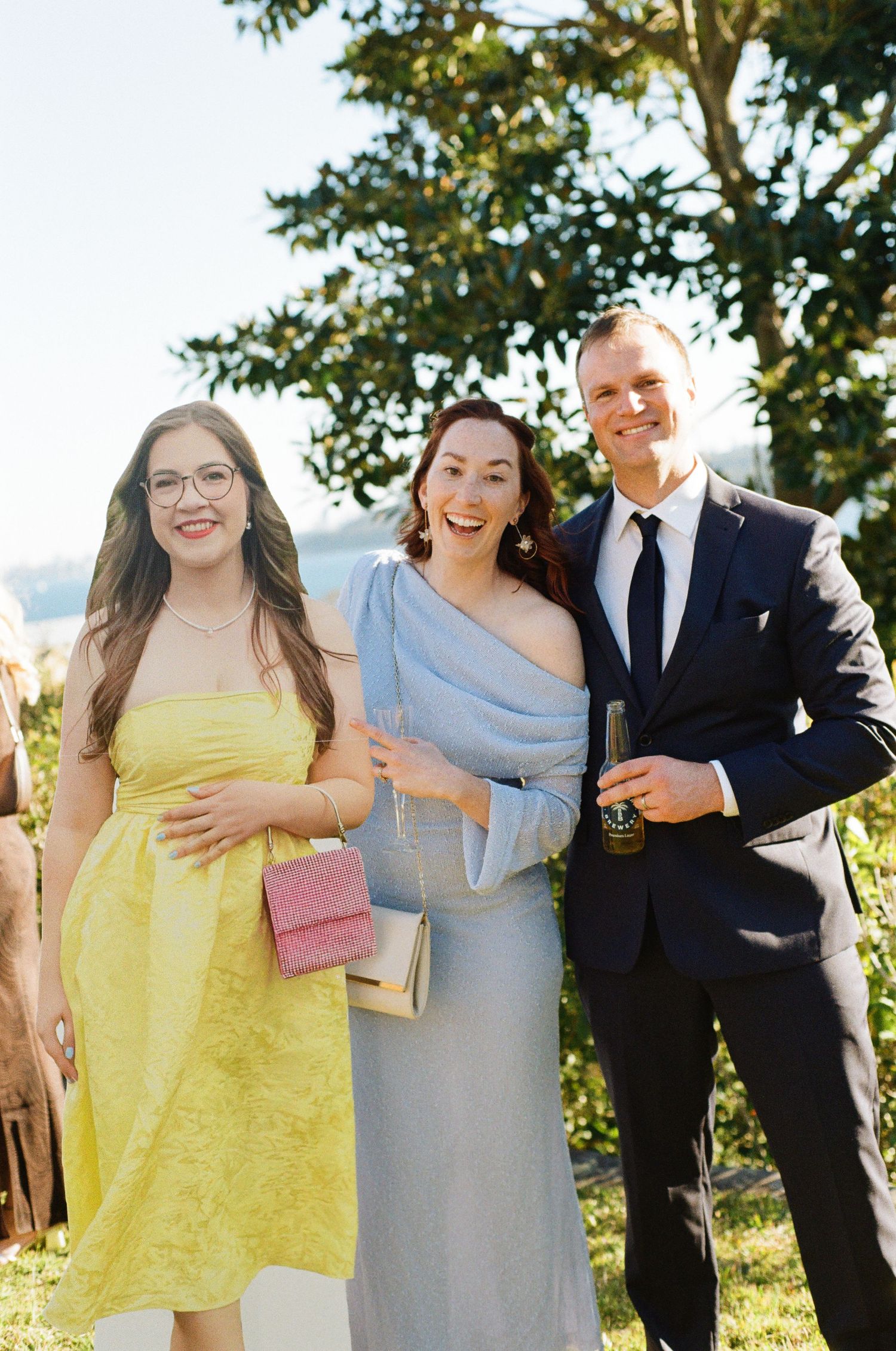 Two women in yellow and blue dresses posing with man in navy suit at outdoor wedding reception.