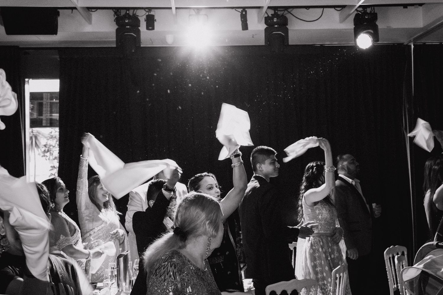 A black and white photo of people waving white napkins in celebration at a festive party venue with stage lighting.