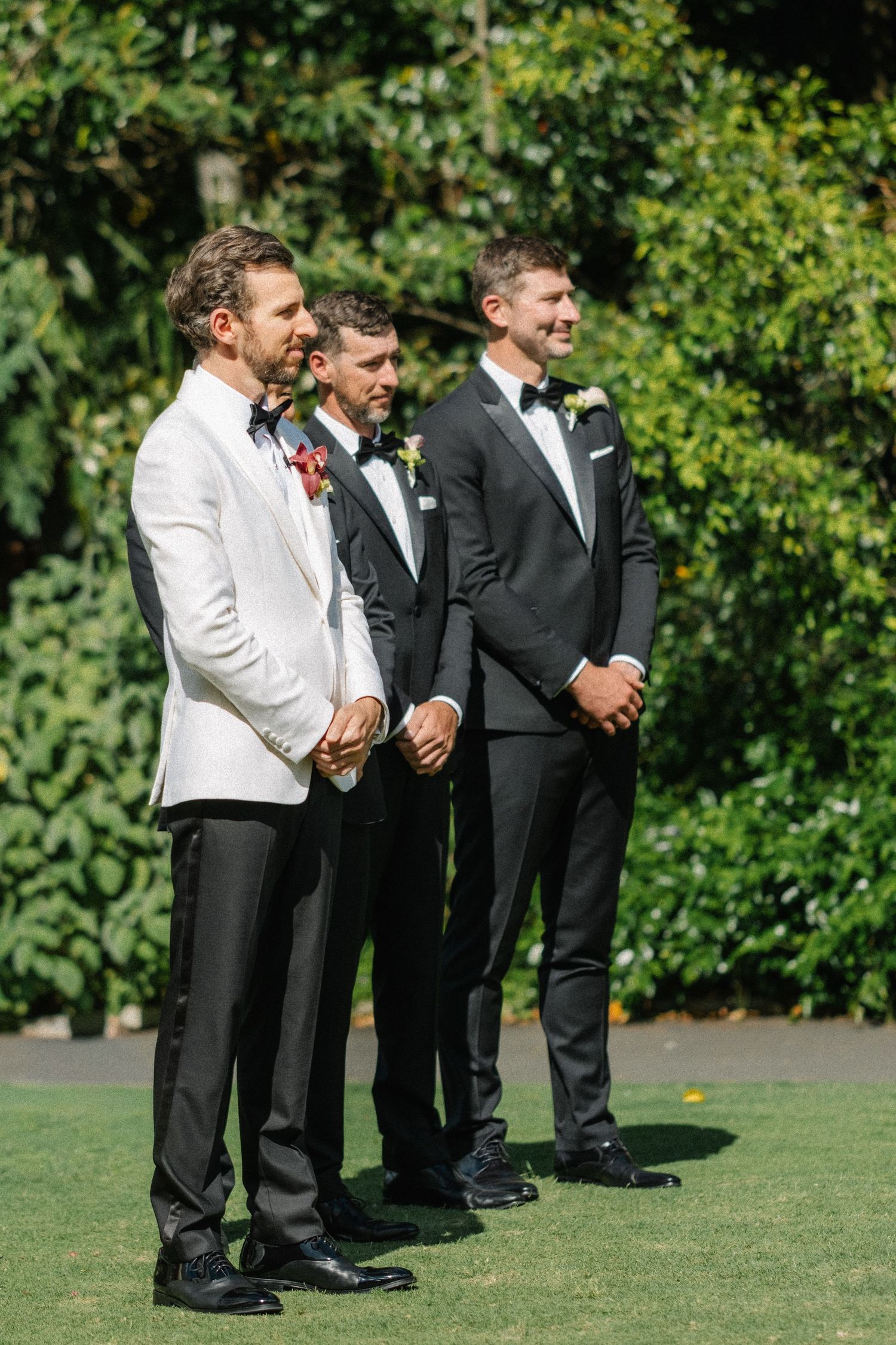 Groomsmen in black and white formal attire stand in a line on green grass during an outdoor wedding ceremony.