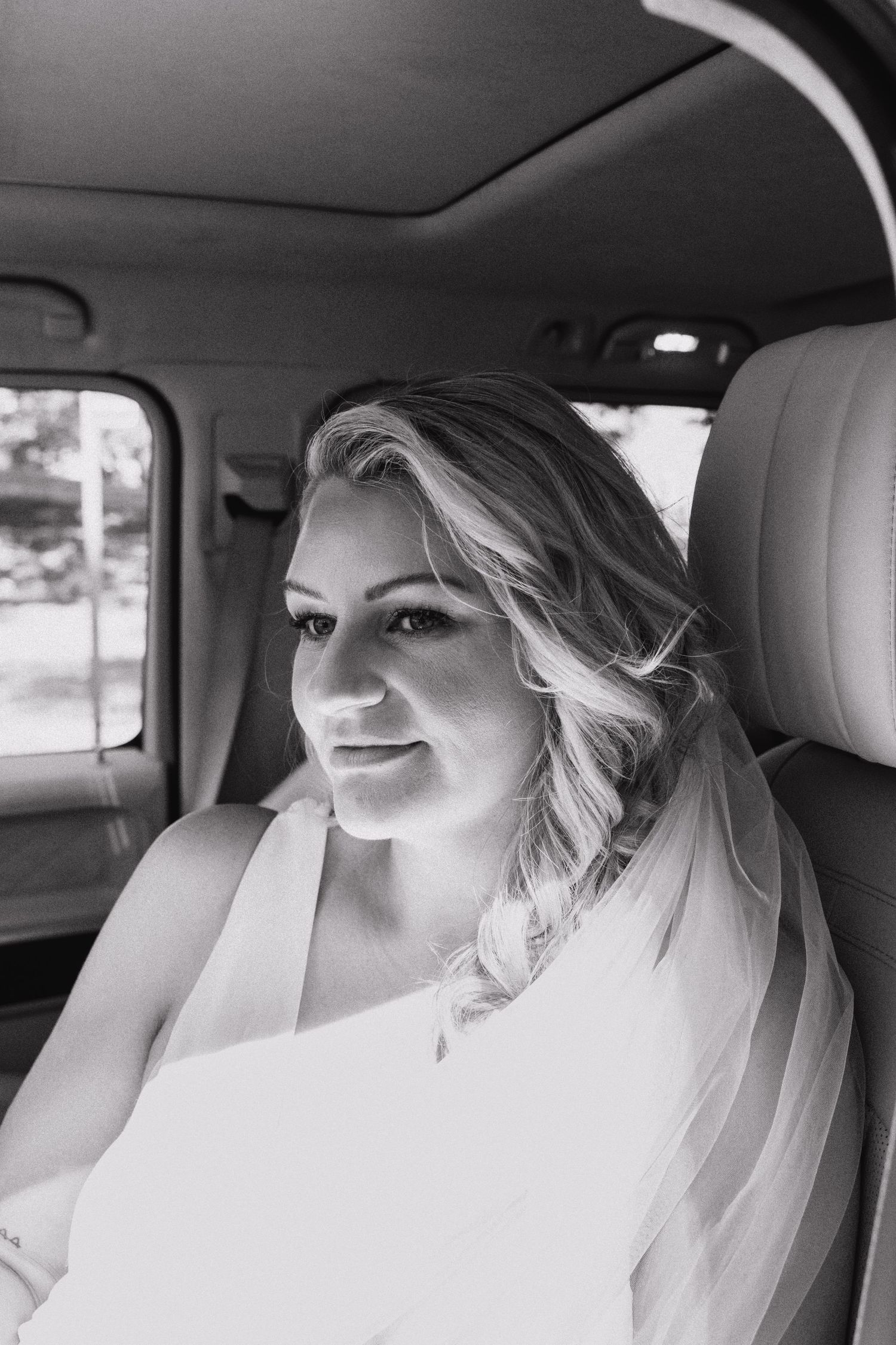 A bride wearing a white wedding dress and veil sits in the backseat of a car on her wedding day.