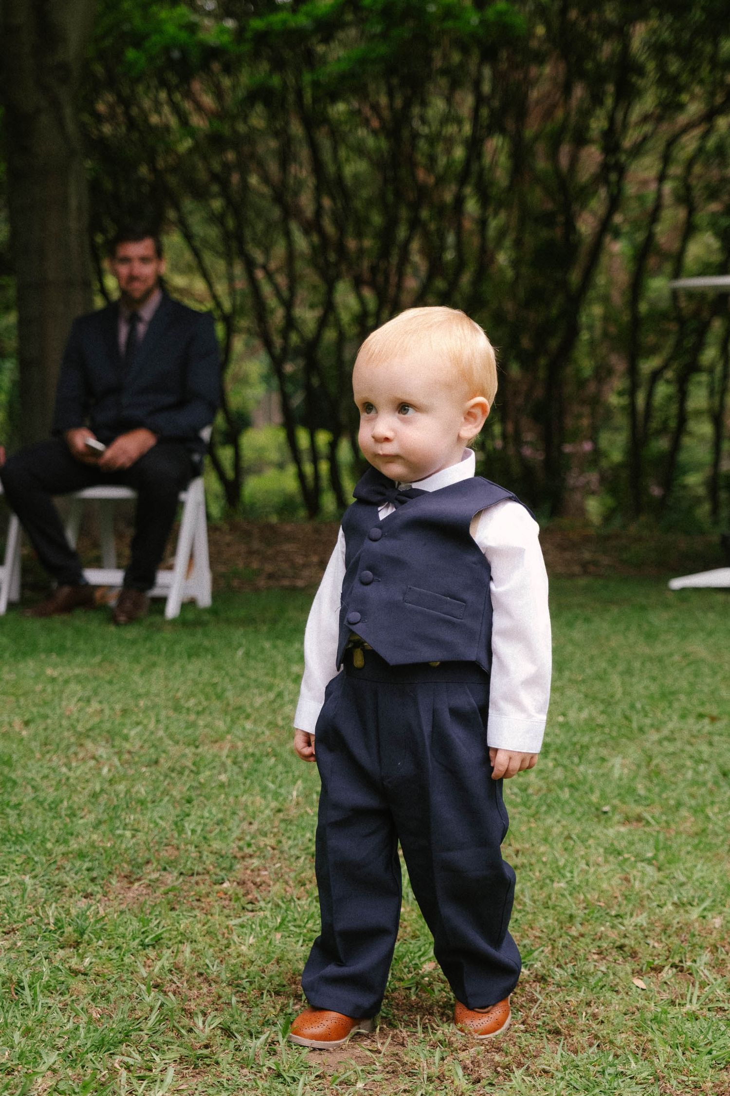 A toddler in a formal navy vest and pants outfit stands on grass during an outdoor event.