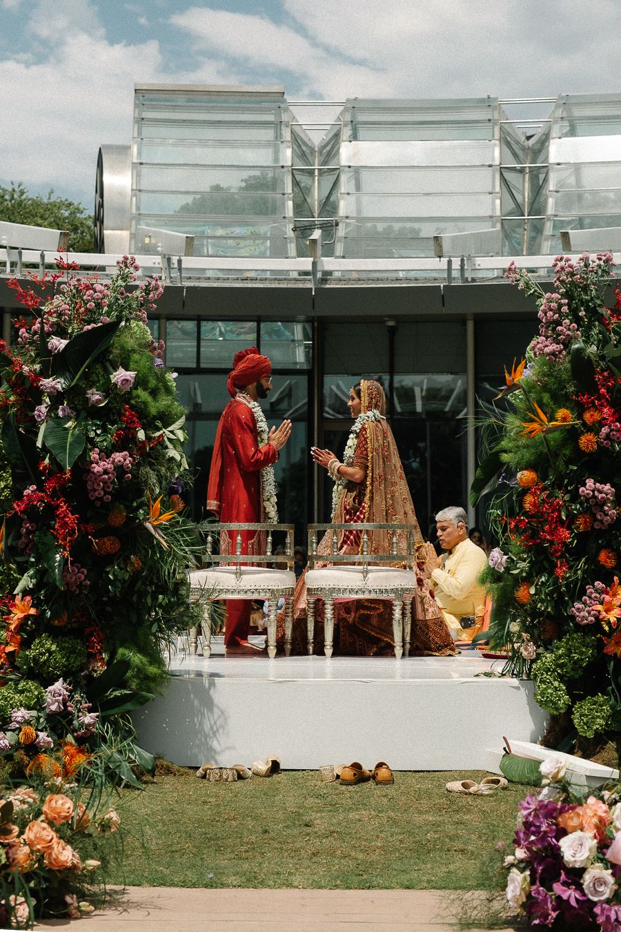 Colorful outdoor wedding ceremony setup with floral arrangements and glass-walled venue in the background.
