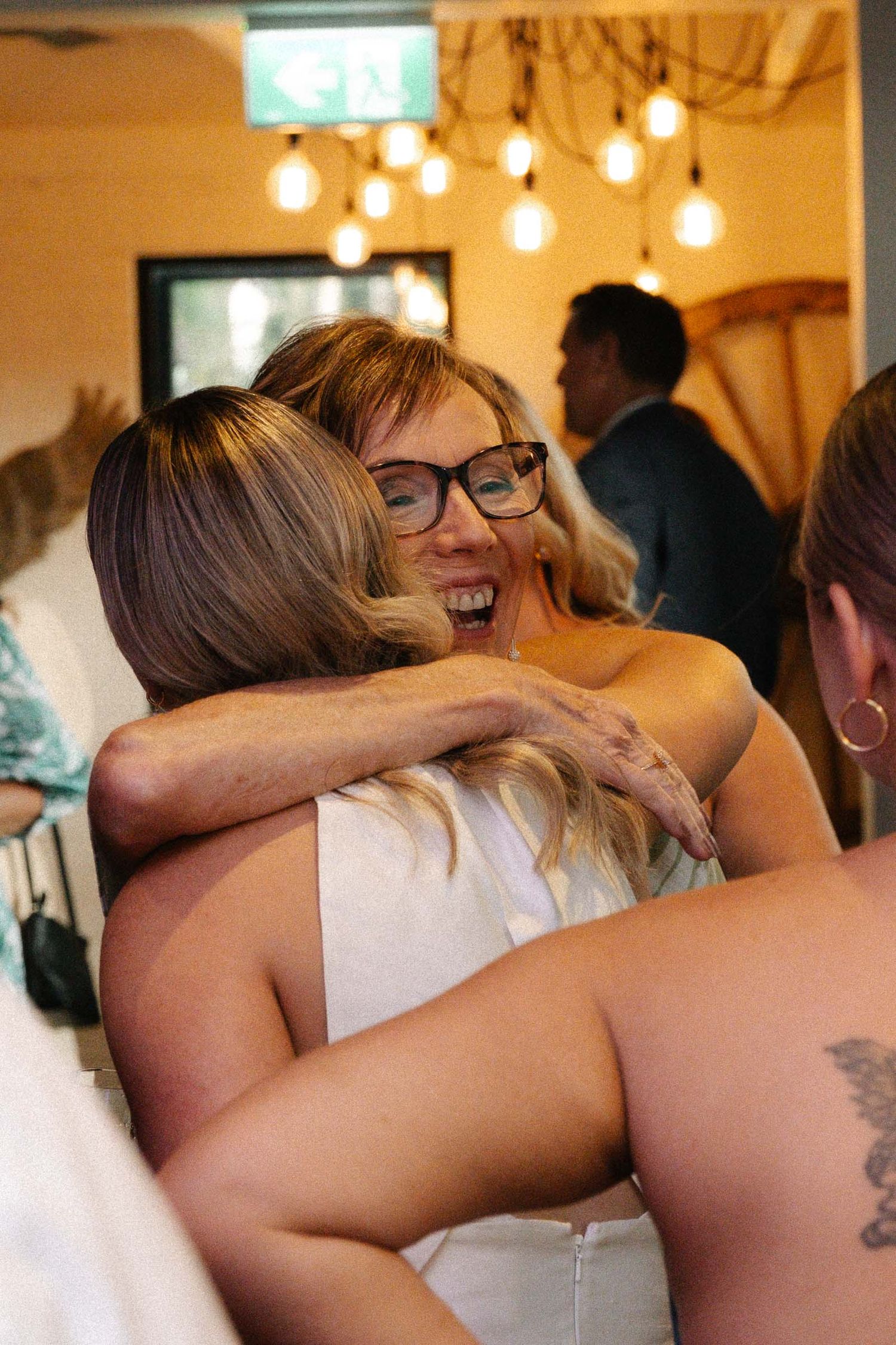 Two people share an emotional embrace at a wedding reception under hanging lights.