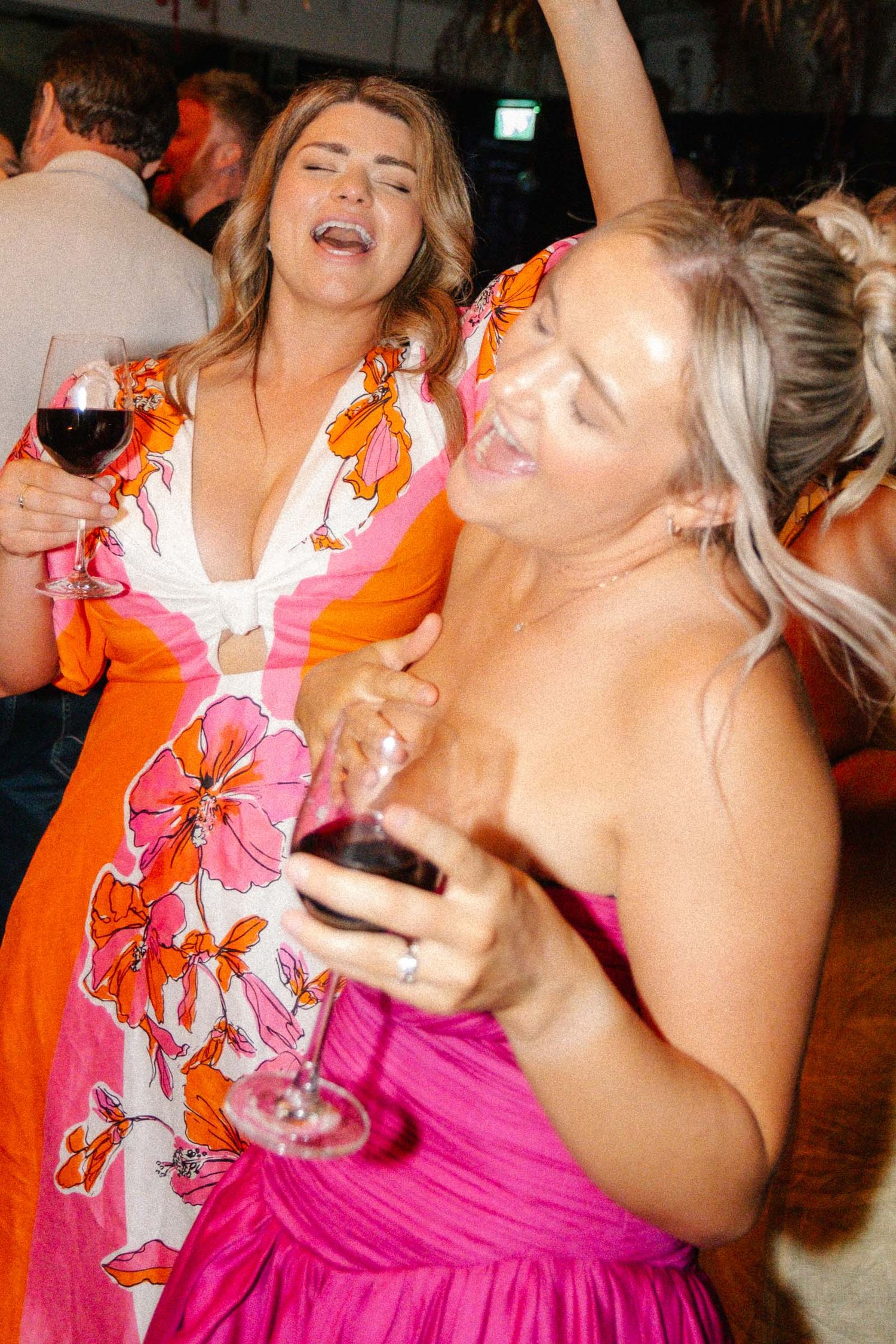 Two women in colorful dresses dancing and laughing while holding glasses of red wine at a party.