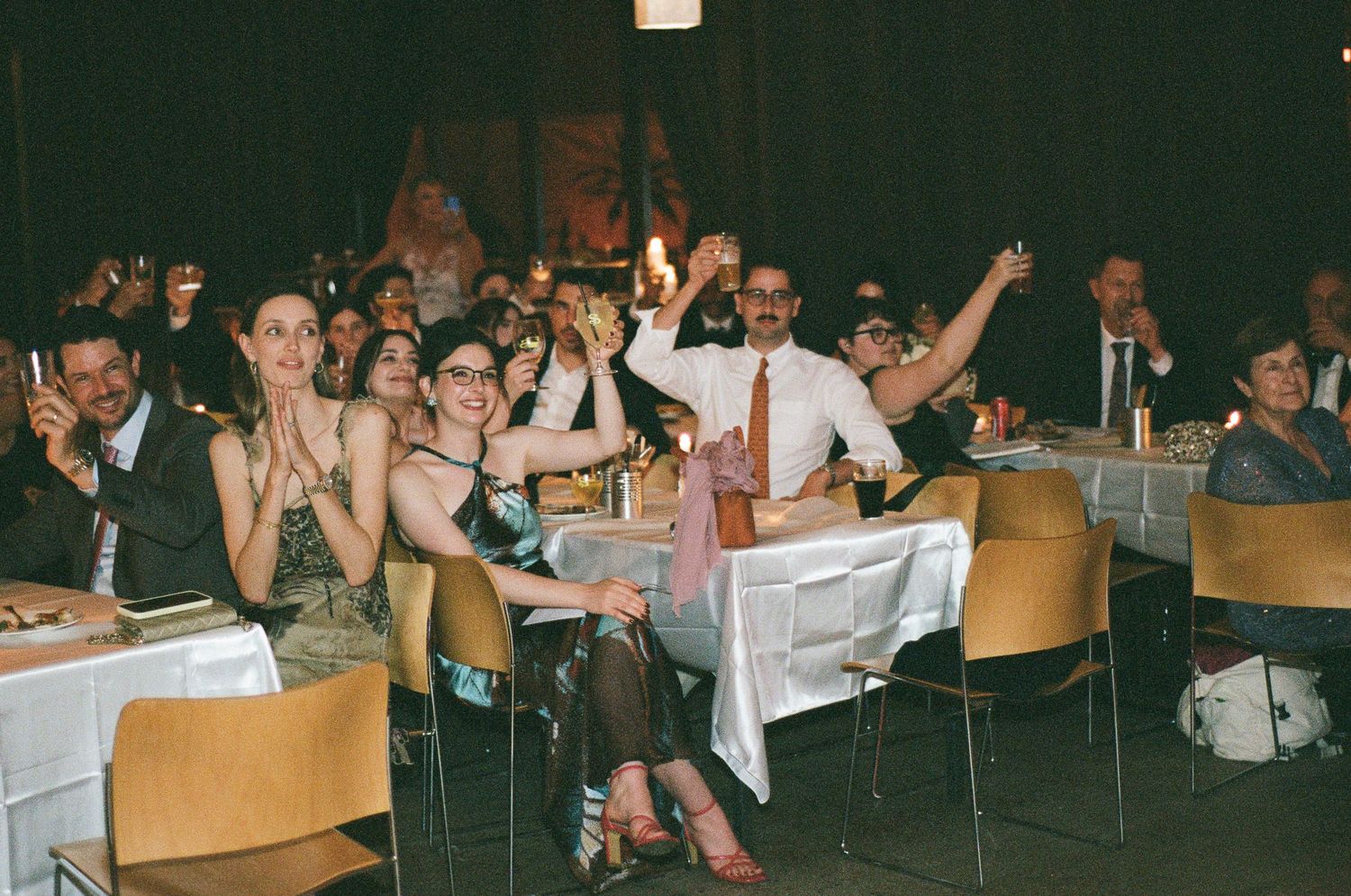 Cheerful guests applaud and raise glasses in a toast at a festive banquet with white-clothed tables in a dimly lit hall.