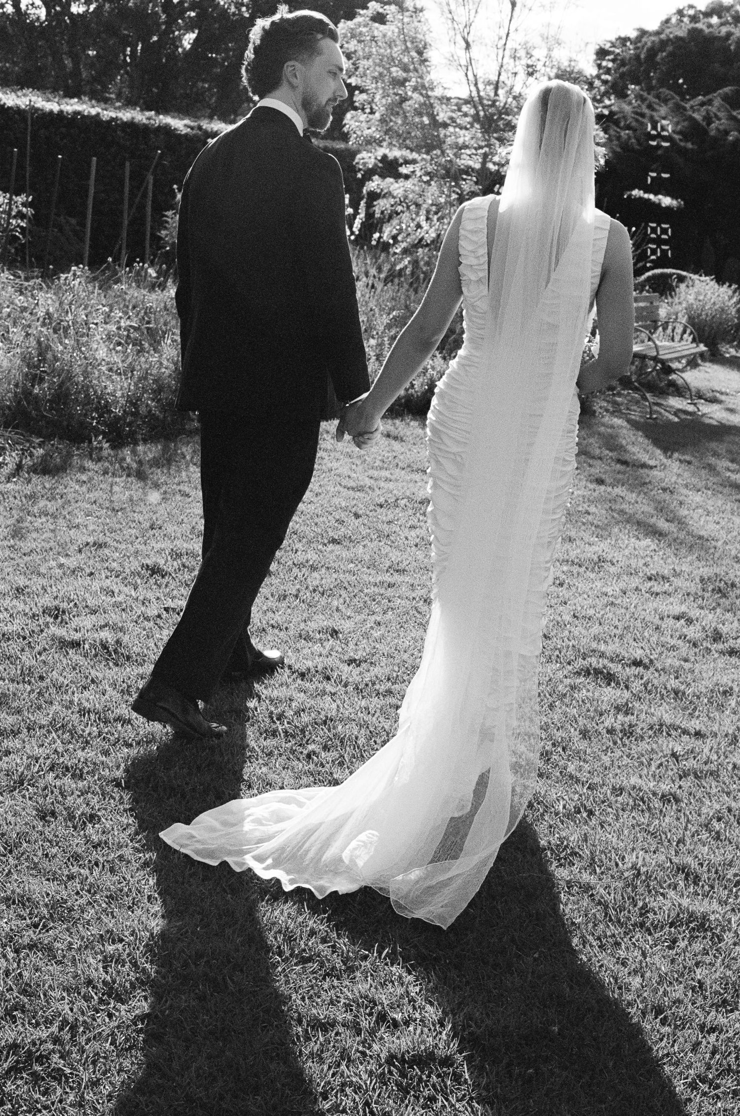 Black and white photo of a bride in a long veil and gown holding hands with a groom in a suit while walking across a sunlit garden lawn.