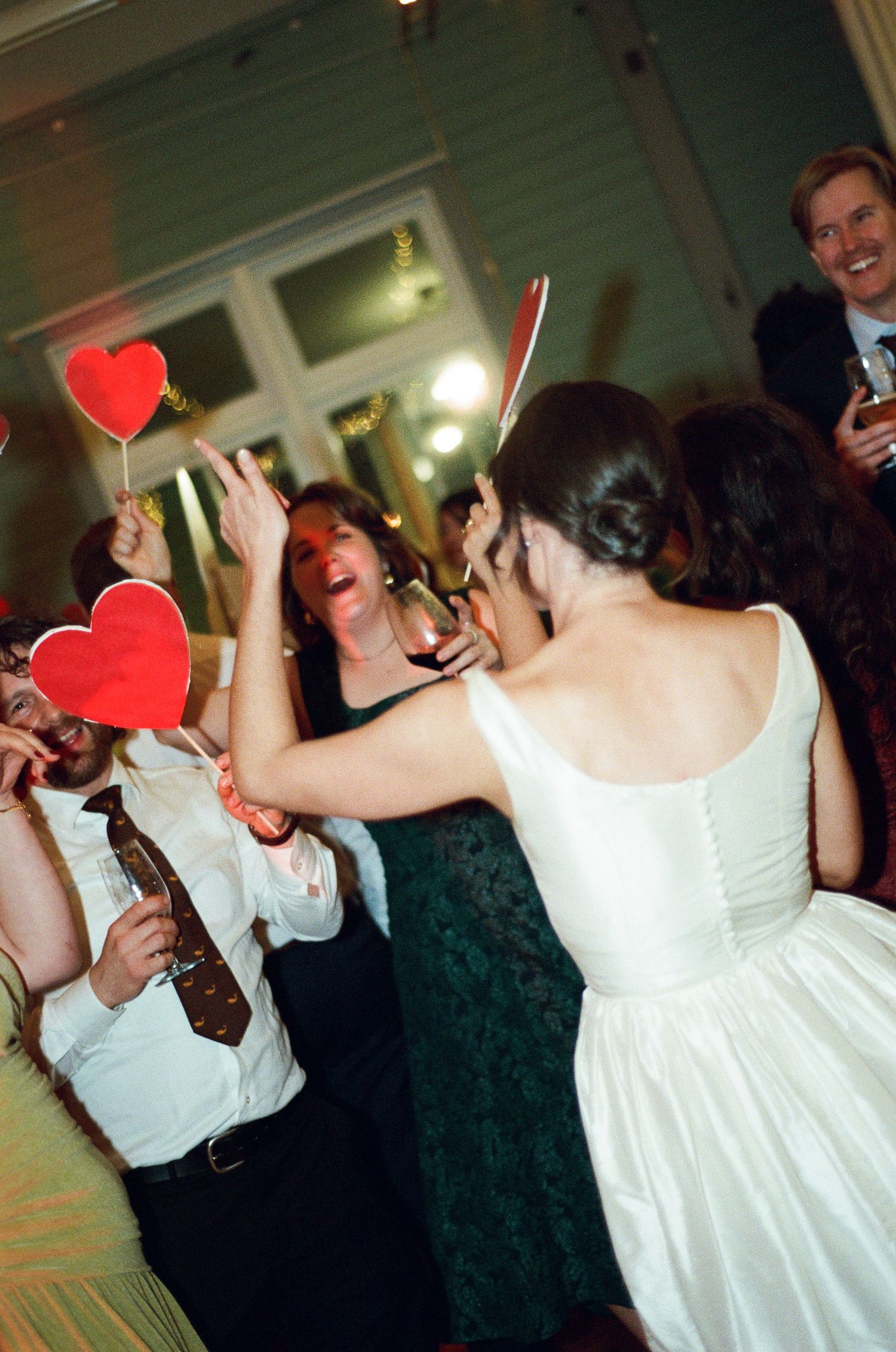 Bride in a white dress with an updo dances joyfully with guests holding red heart signs and wine glasses at a lively wedding reception.