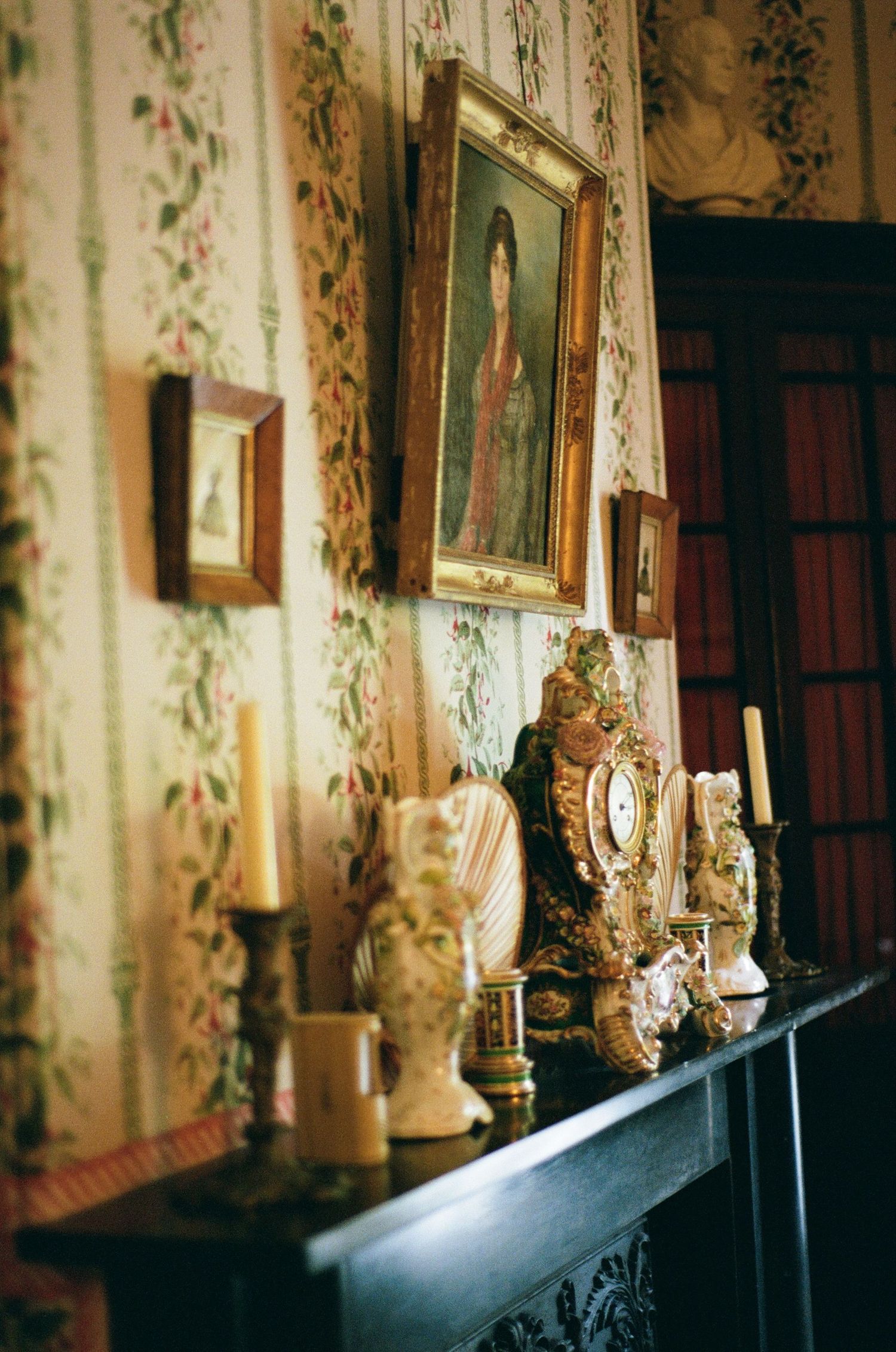 Ornate antique candelabras and decorative items line a dark wooden shelf against floral wallpaper in a historic interior.