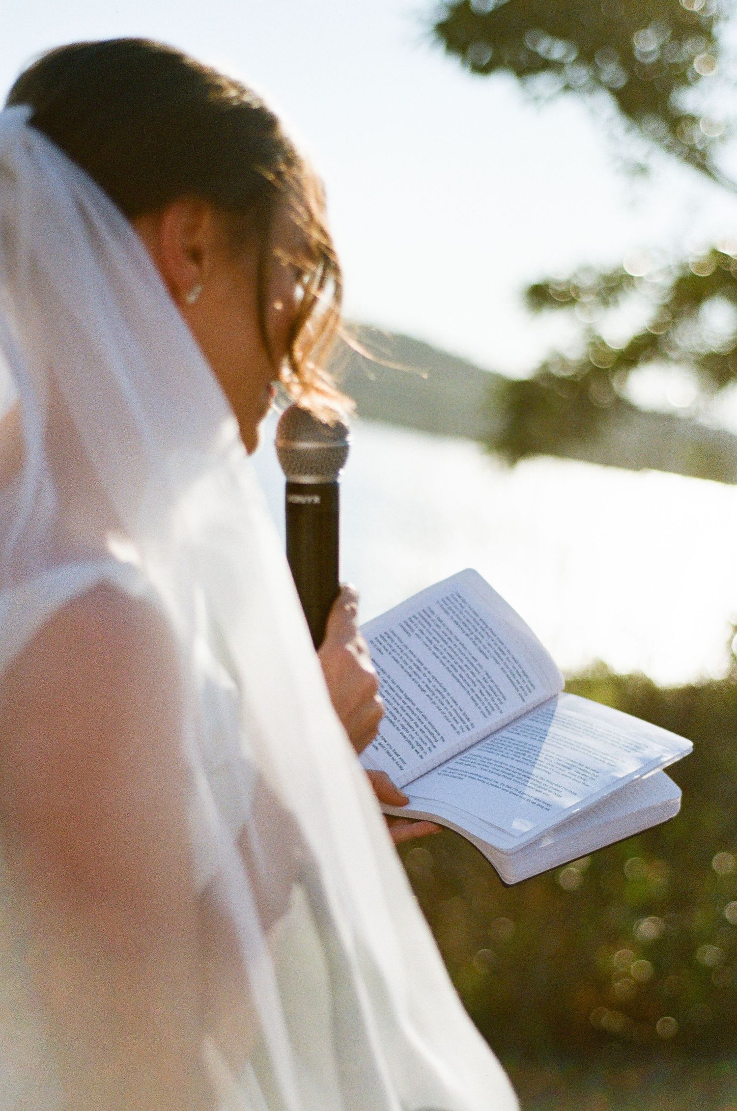 Close-up of bride in veil reading from book while holding microphone at wedding ceremony.