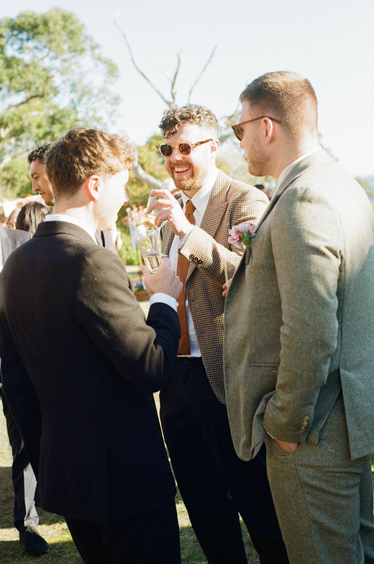 Groomsmen in suits laughing and toasting drinks outdoors at a sunny wedding celebration.