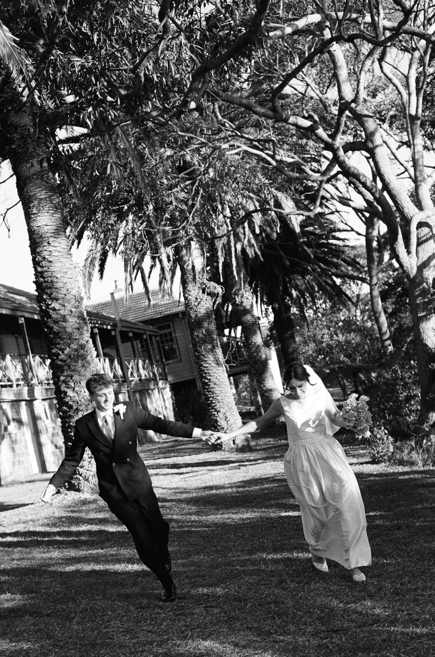 Bride and groom hold hands and run joyfully across a sunlit lawn beneath tall trees in a black and white wedding photo.