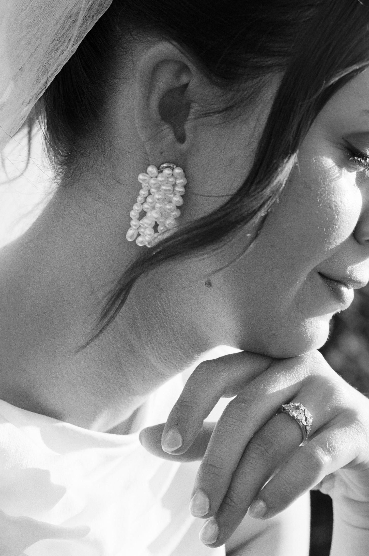 Black and white close-up of a bride in a veil wearing pearl cluster earrings and a diamond ring while resting her chin on her hand.