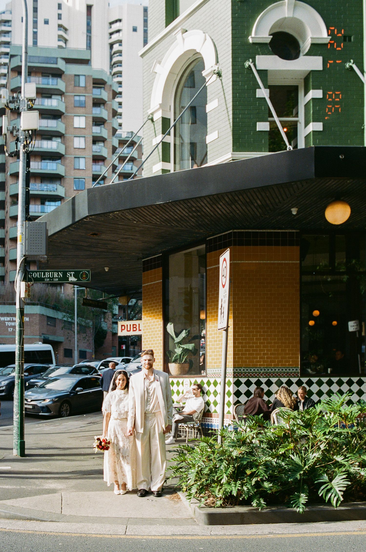 Bride and groom in cream wedding attire pose on a city street corner outside a green and yellow tiled pub with apartment towers behind.
