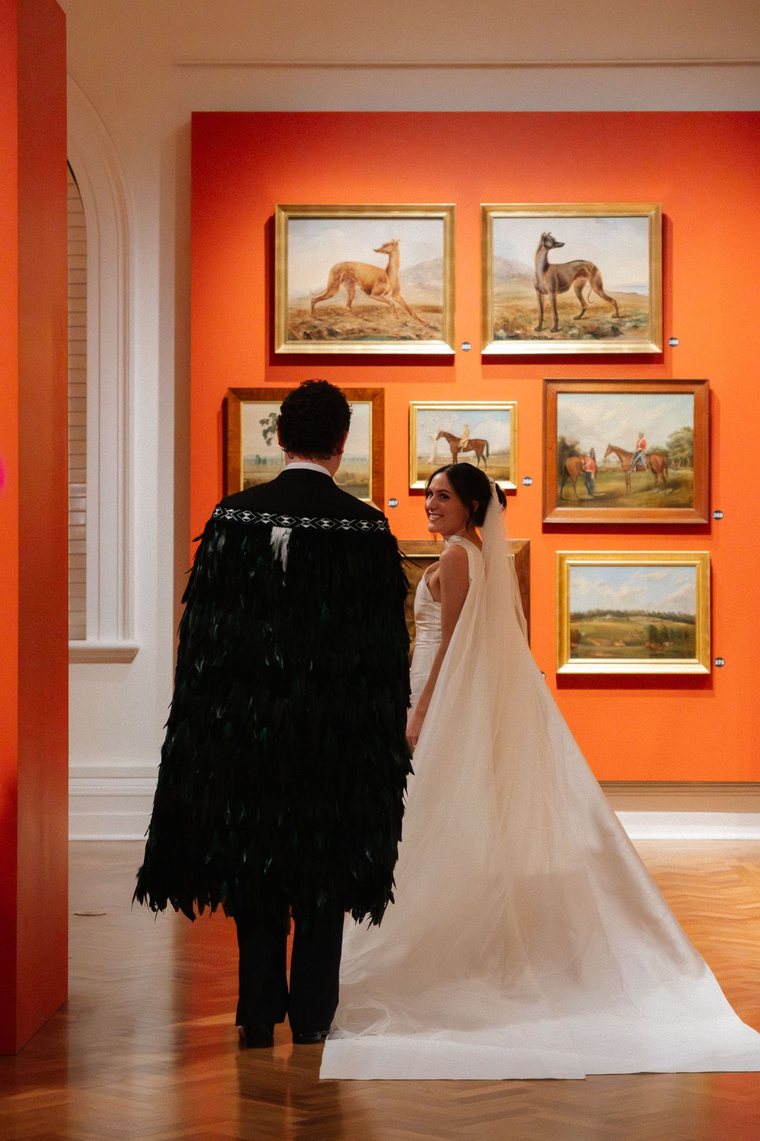 A couple in wedding attire views artwork in an art gallery with orange walls displaying paintings of horses.