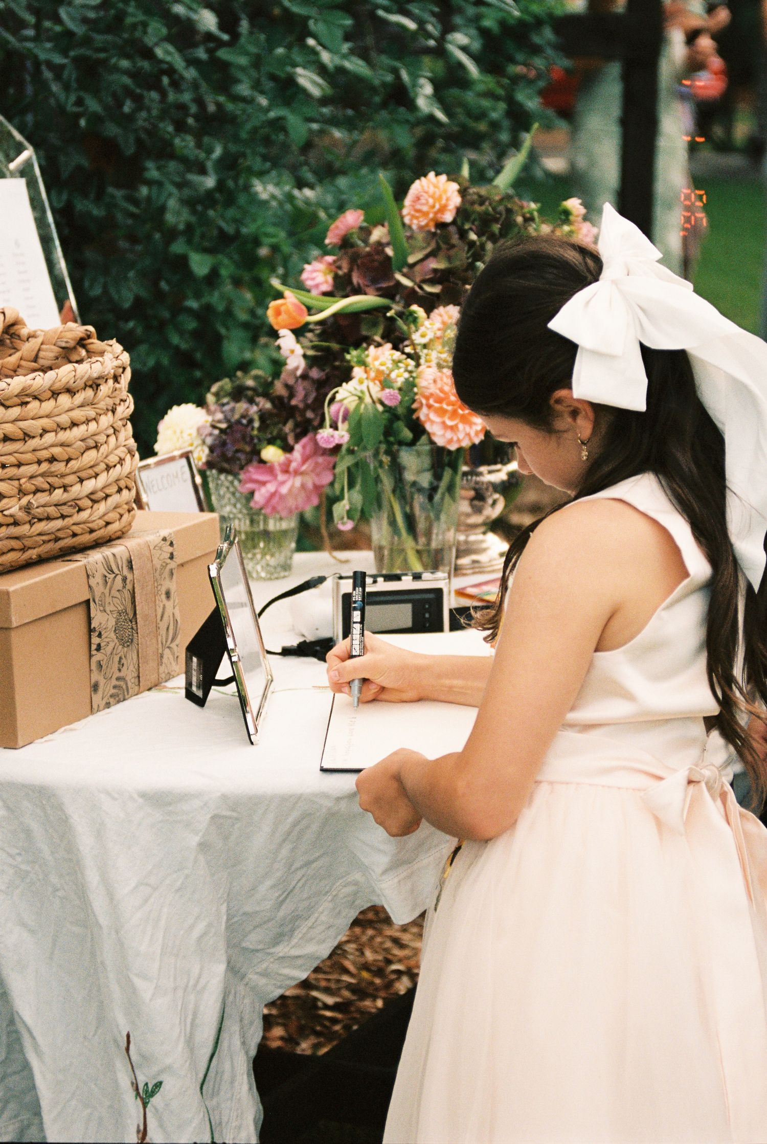 Girl in a blush dress with a large white bow signs a guestbook at a wedding table decorated with colorful flowers and a woven basket.