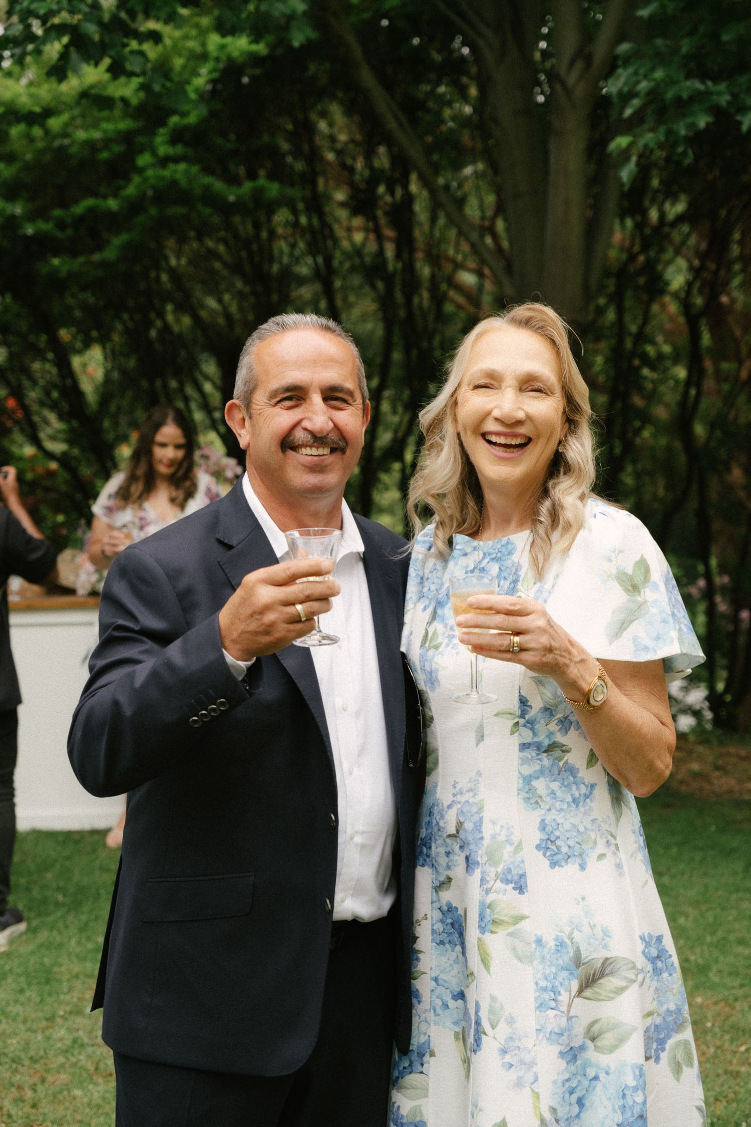 A couple shares a celebratory toast outdoors while wearing formal attire at a garden event.