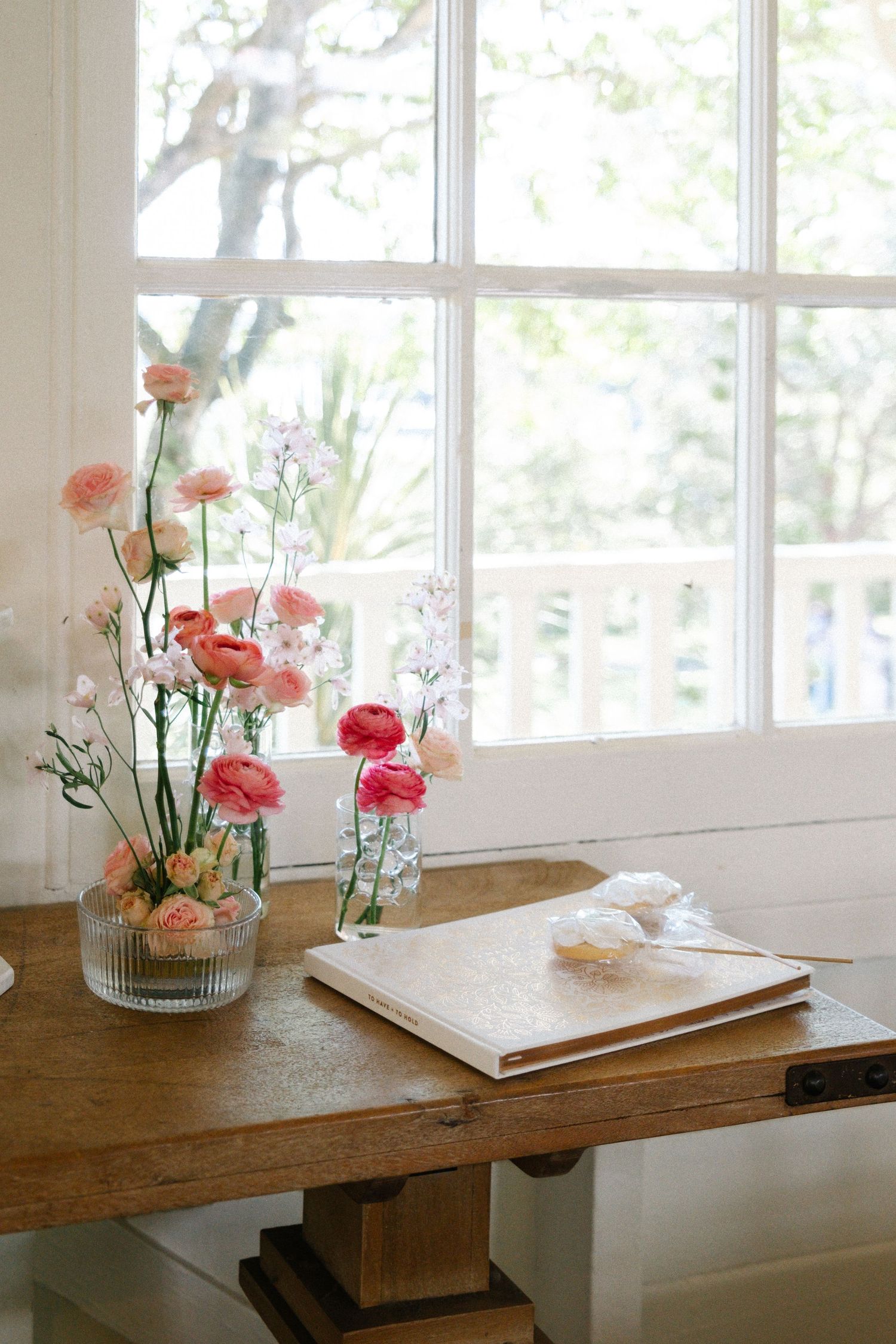 Pink carnations in a glass vase sit on a rustic wooden table by a bright window with natural light streaming in.