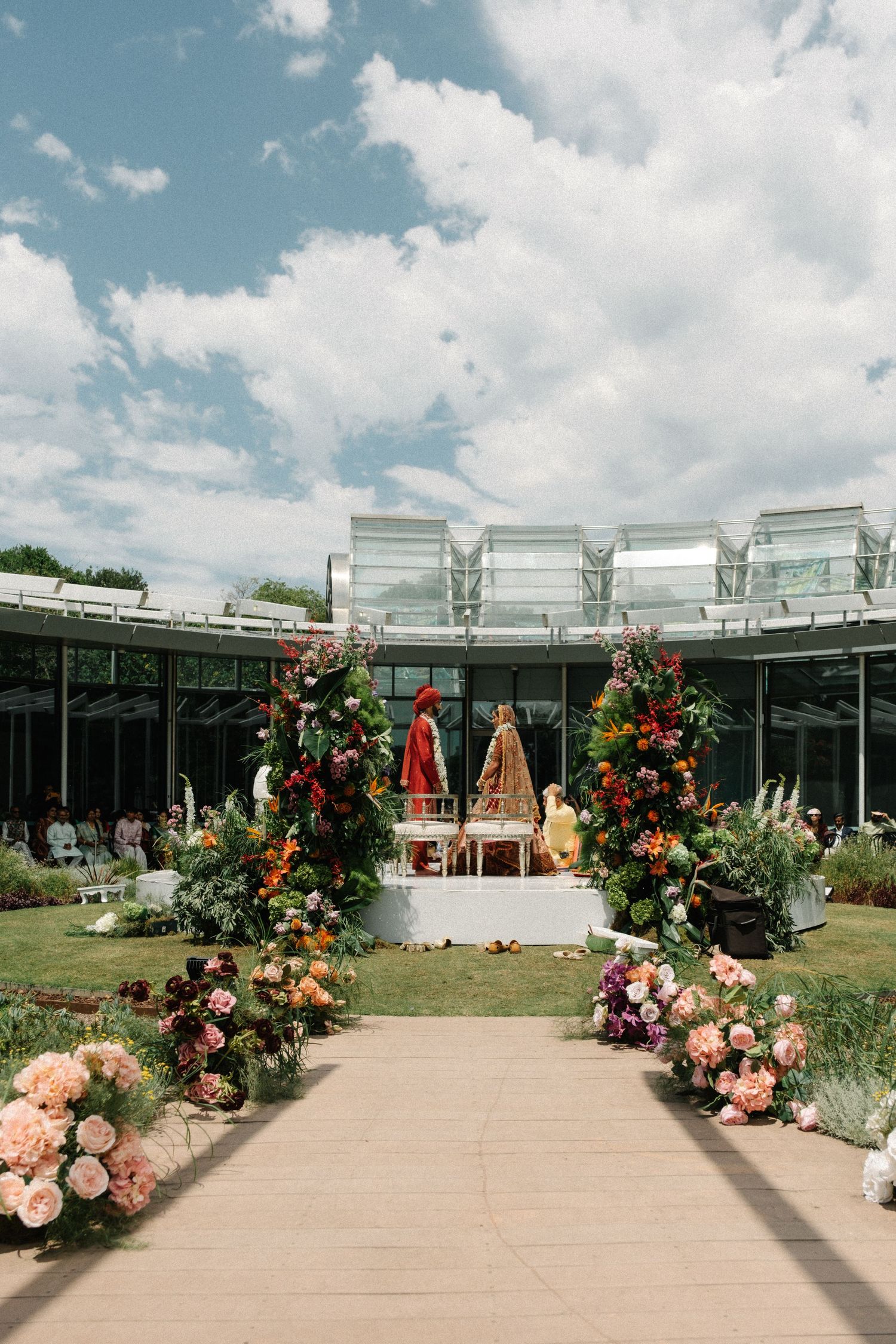 Outdoor wedding venue setup with glass greenhouse structure and floral arrangements along a paved walkway under blue skies.