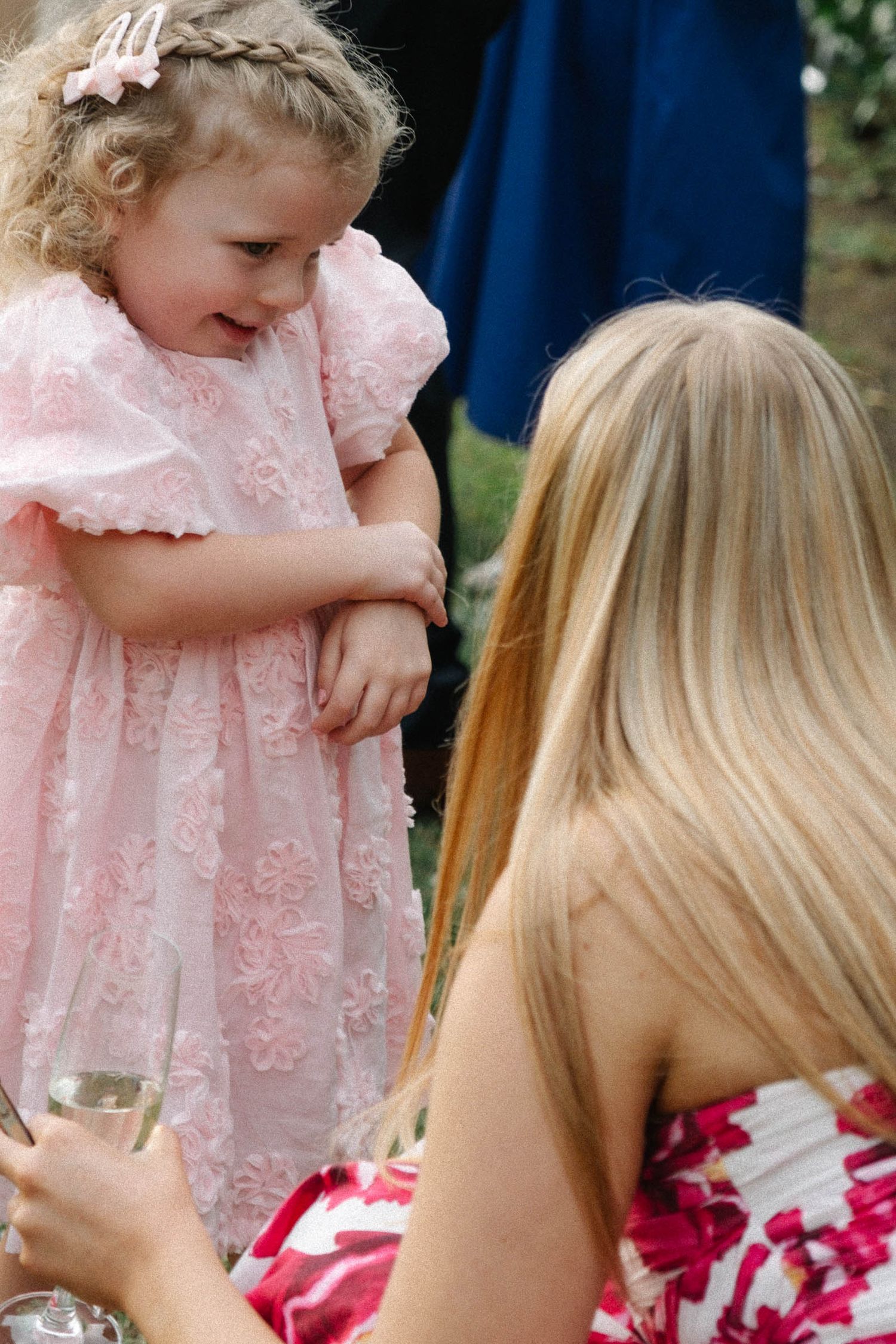 A person in a pink lace dress interacts with someone wearing a floral dress outdoors.