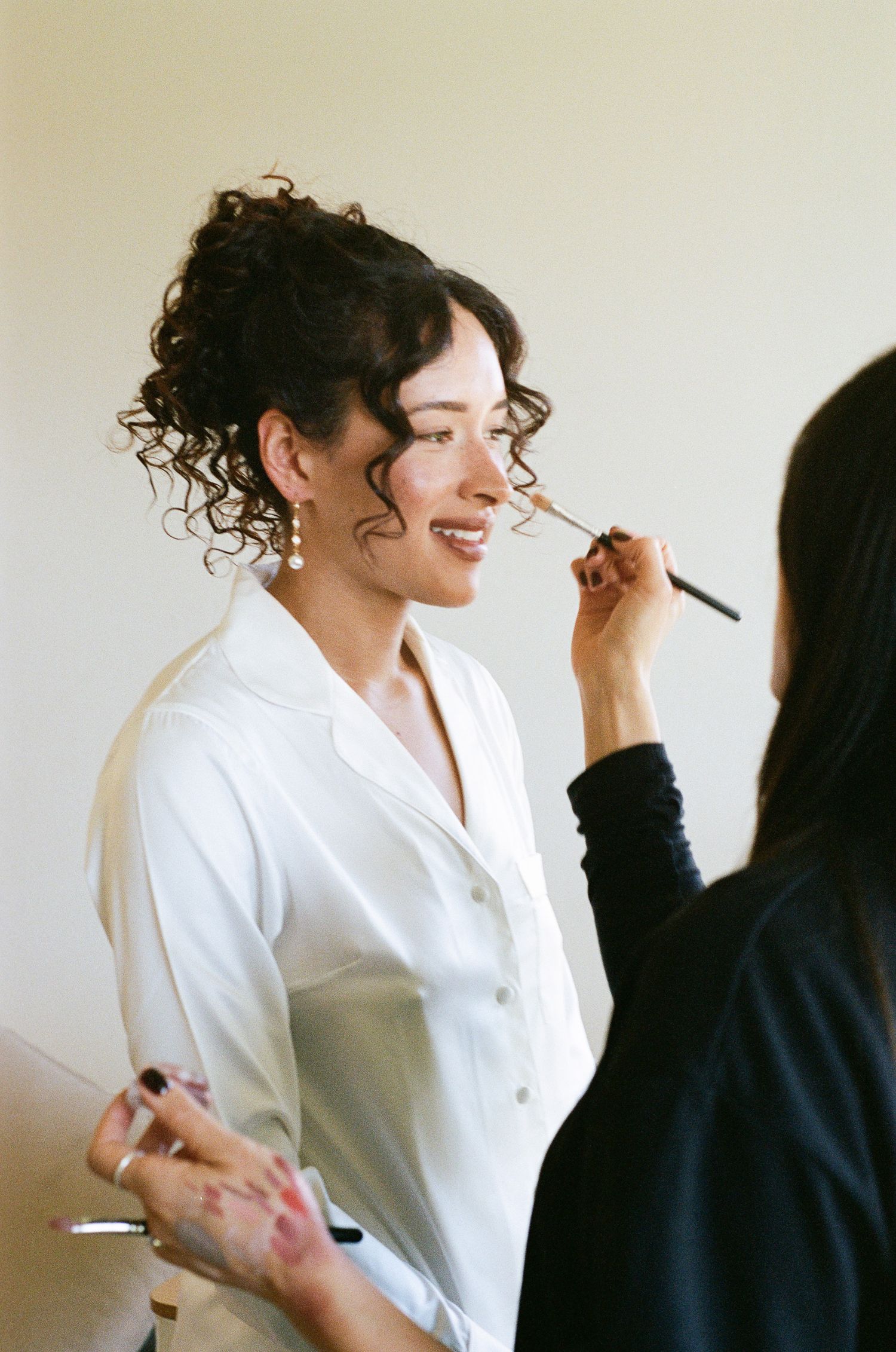 A smiling woman with curly updo hair and a white satin blouse has makeup applied with a brush by a makeup artist.