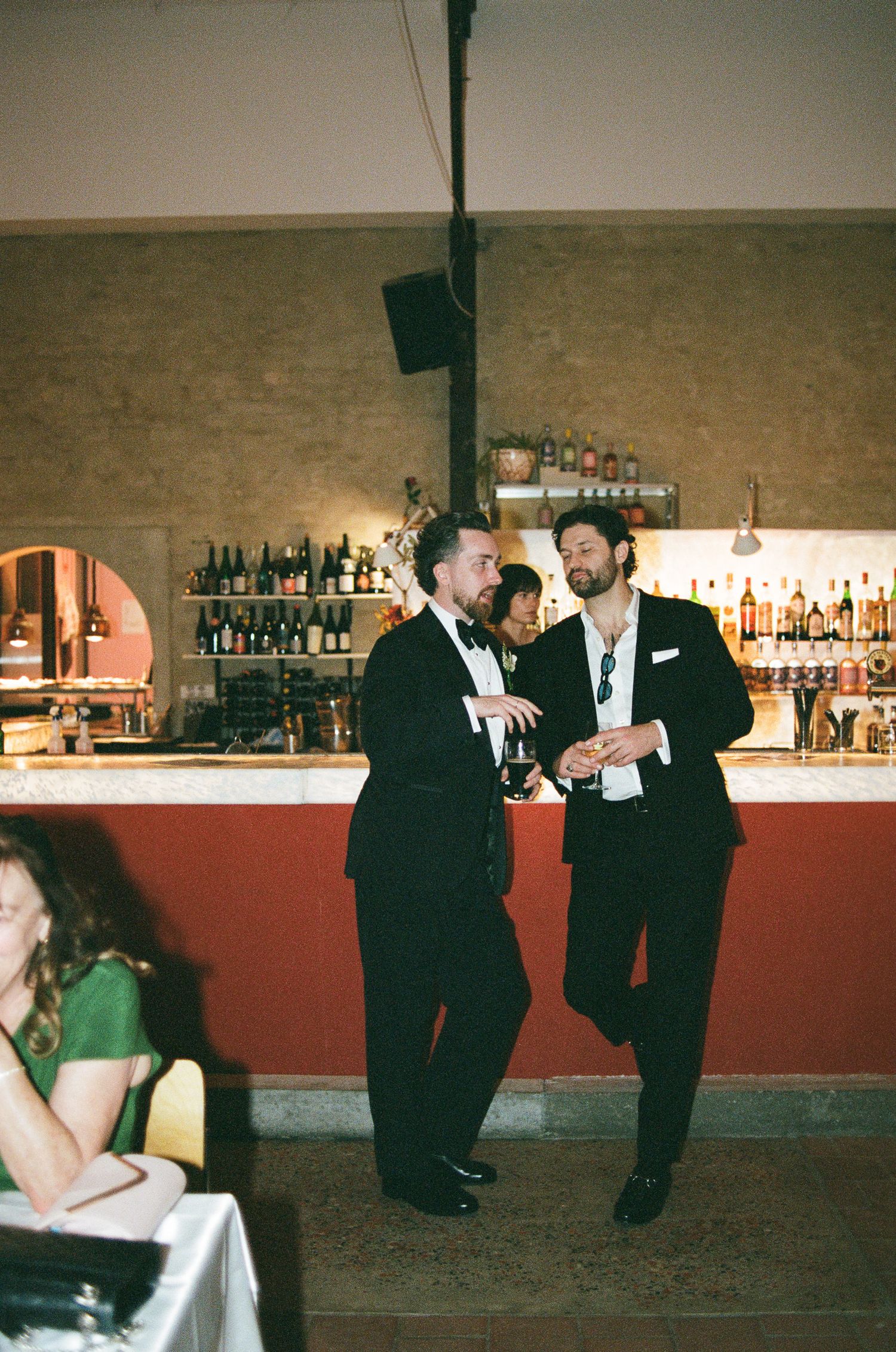 Two men in black tuxedos stand talking in front of a bar lined with liquor bottles while a woman in green sits at a table nearby.