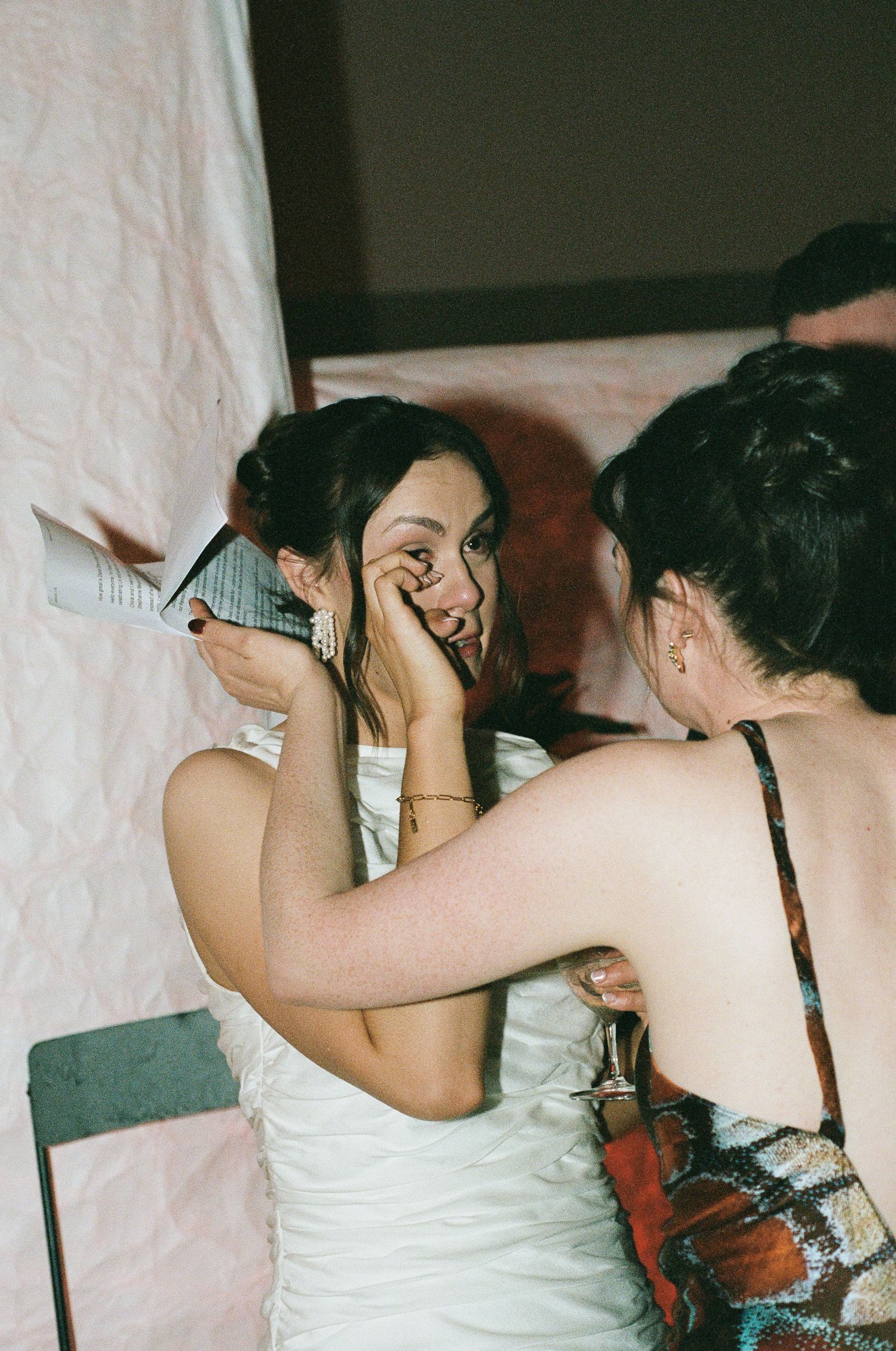 A woman in a white satin dress wipes her eyes while another woman in a patterned top comforts her at an emotional celebration.