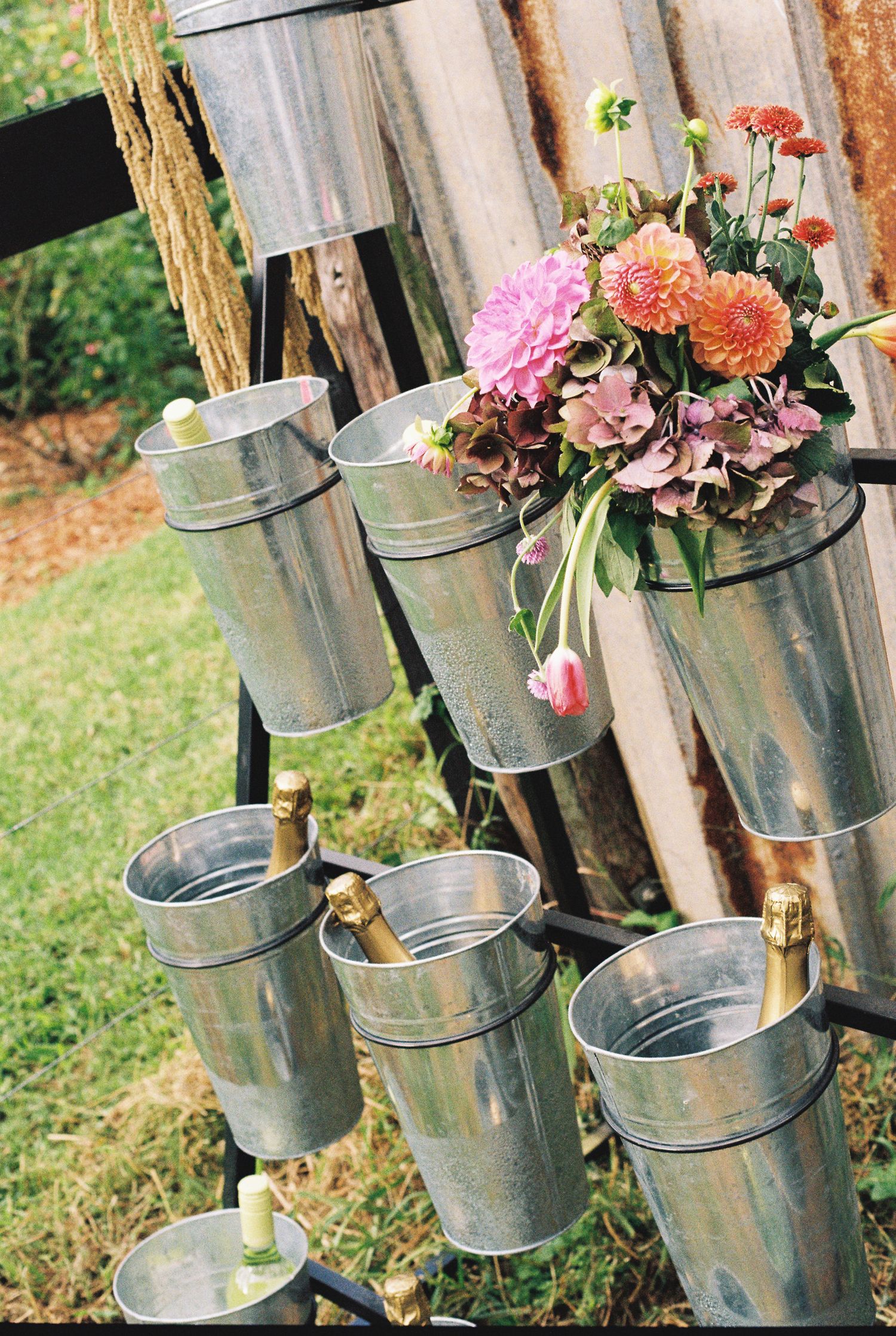 Galvanized metal buckets hung on a rustic wooden stand hold champagne bottles and a bouquet of pink and orange flowers outdoors.