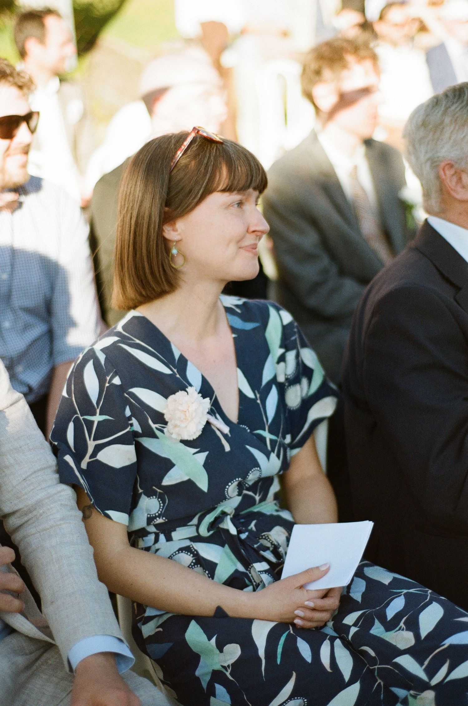 Wedding guest in floral navy dress holds program while seated at outdoor ceremony.