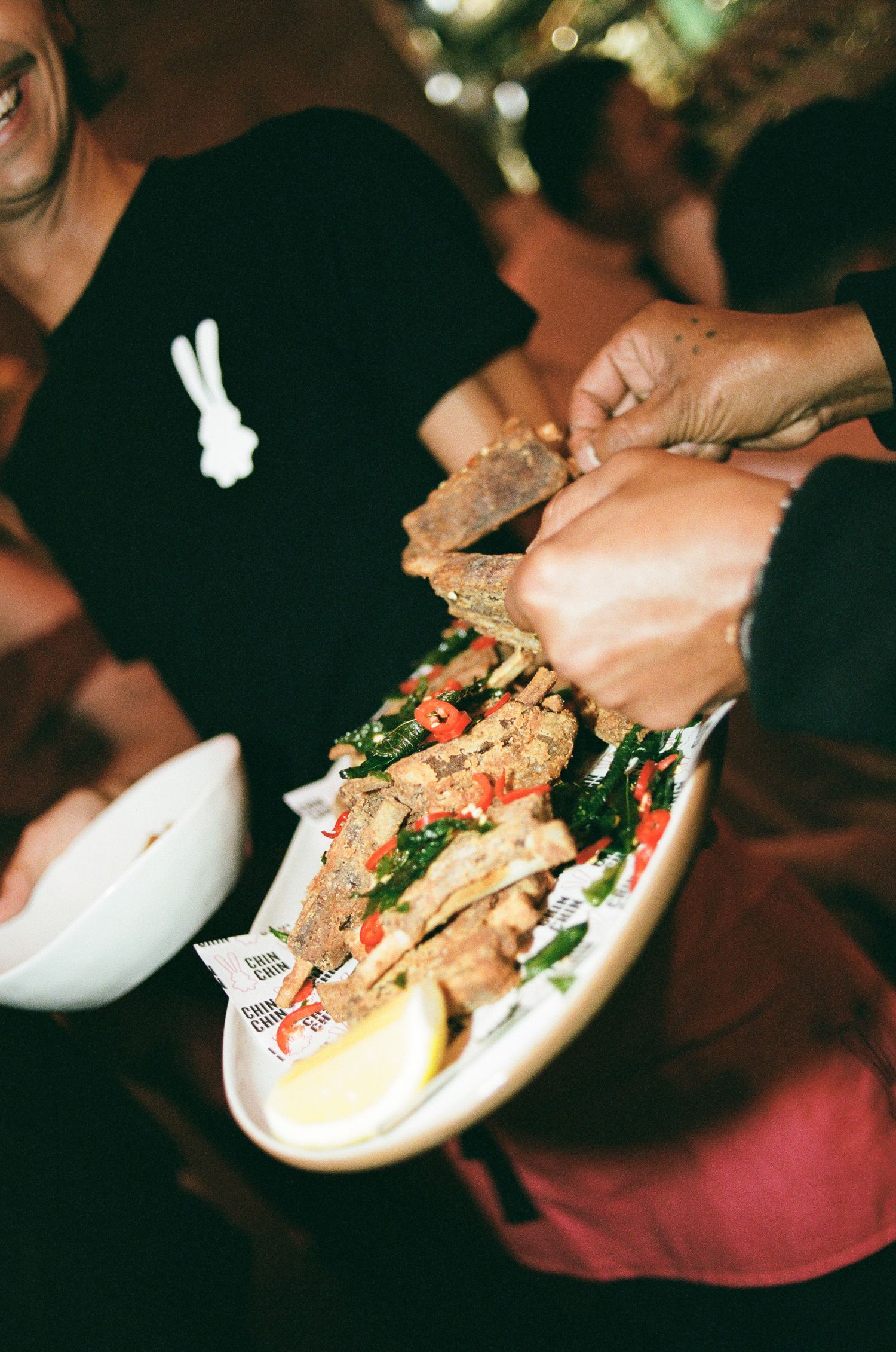 Hands pick up a piece of crispy fried food from a plate with chili peppers, fresh greens and a lemon wedge as a smiling person looks on.