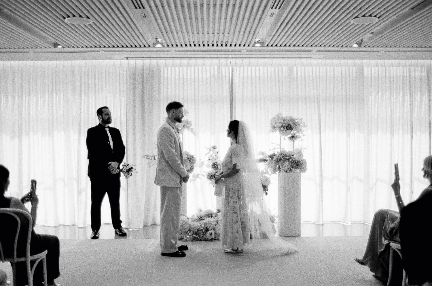 Black and white photo of a bride and groom exchanging vows at an indoor wedding ceremony with floral arrangements and sheer curtains.