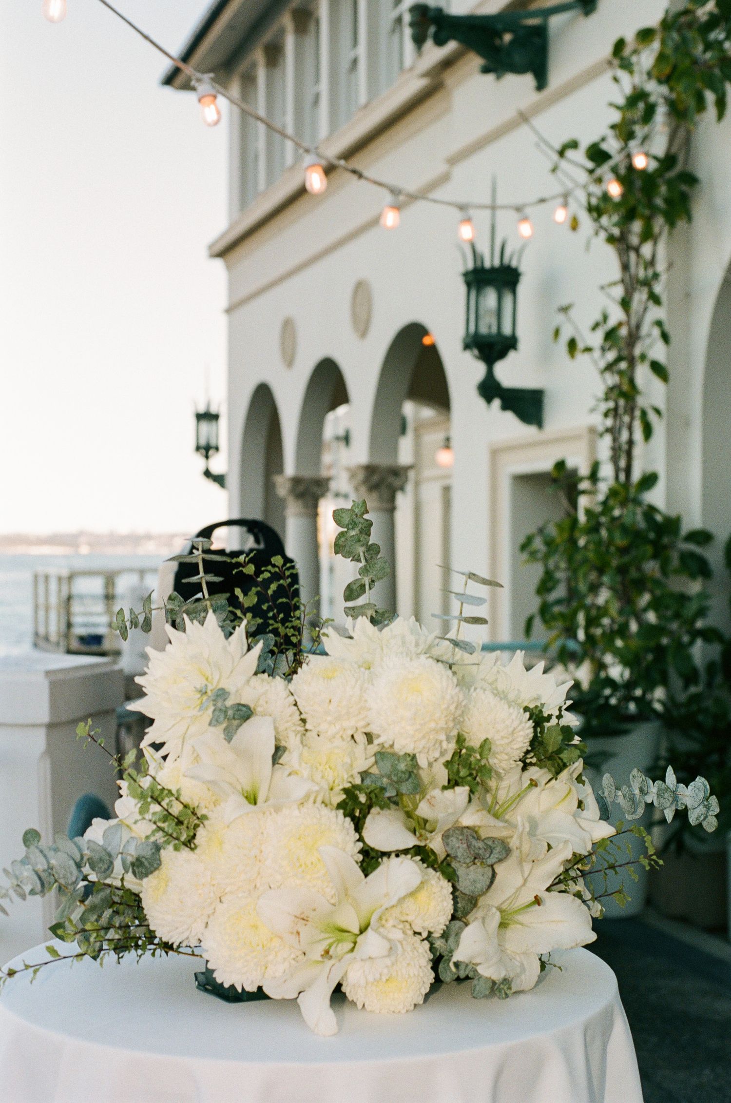 White floral arrangement with eucalyptus on a draped table beside an arched waterfront building with string lights.