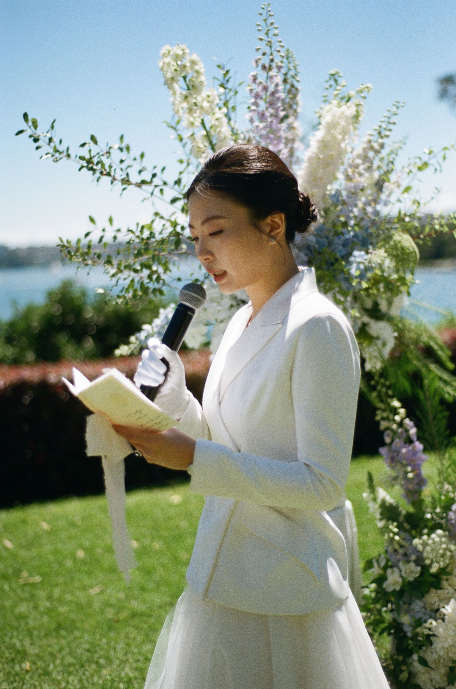 Bride in a white blazer and gloves reads her vows into a microphone at an outdoor lakeside wedding ceremony beside lush floral arrangements.