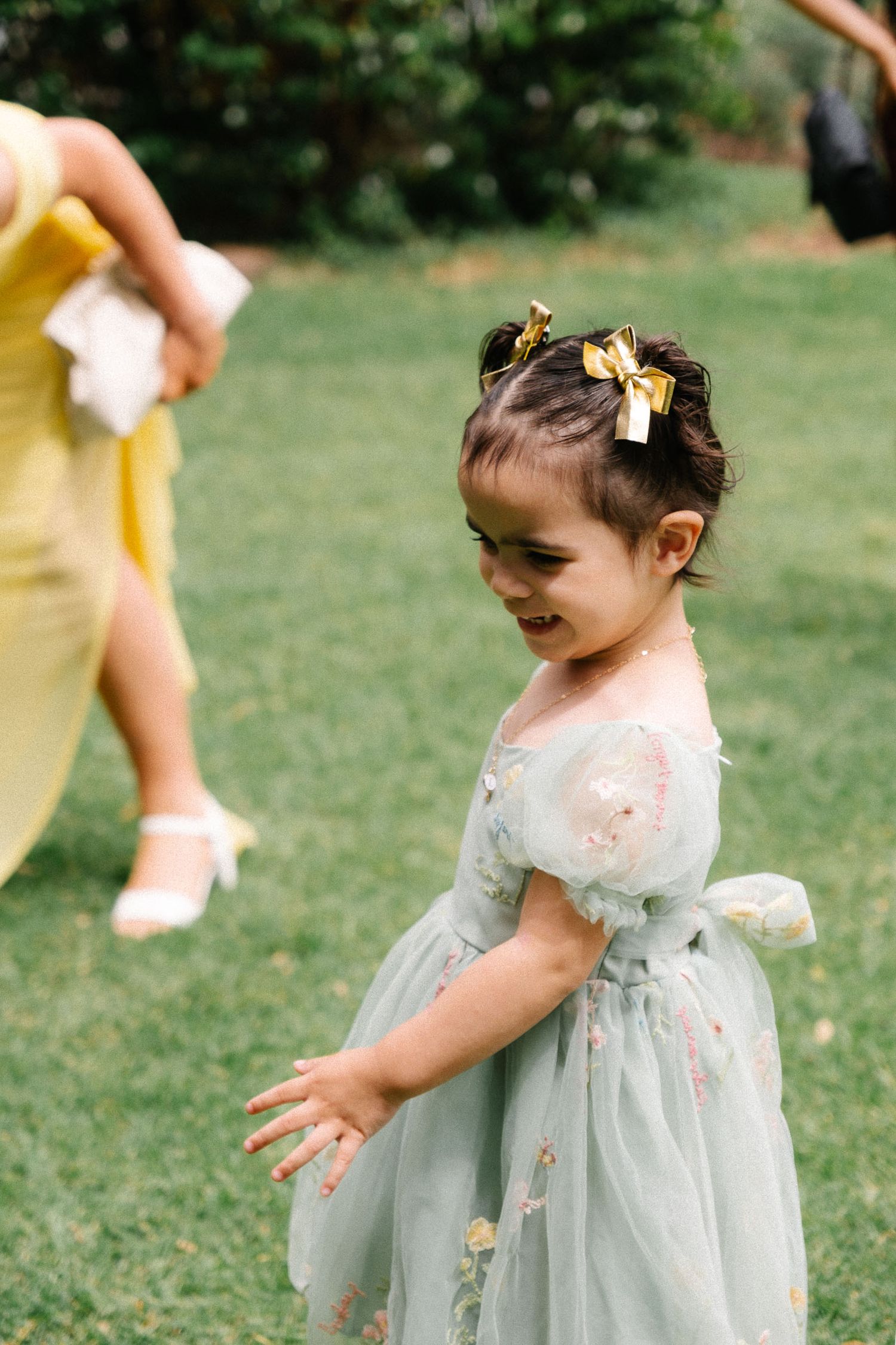 A child in a light blue floral dress plays joyfully on a grassy lawn on a sunny day.