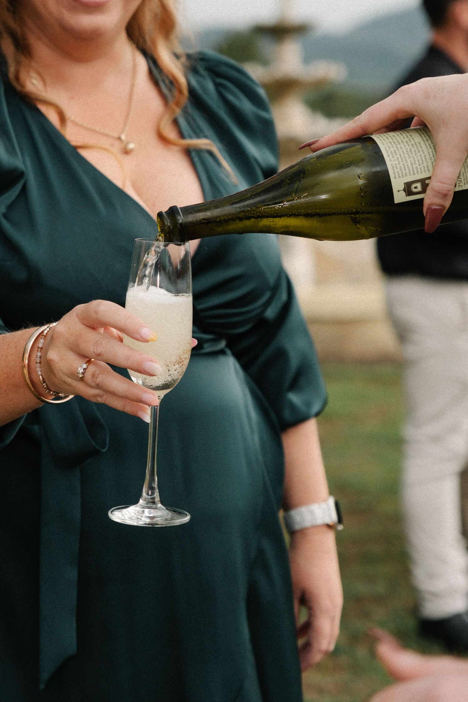 Someone wearing a dark green dress pours champagne into a glass while wearing jewelry and bracelets at an outdoor event.